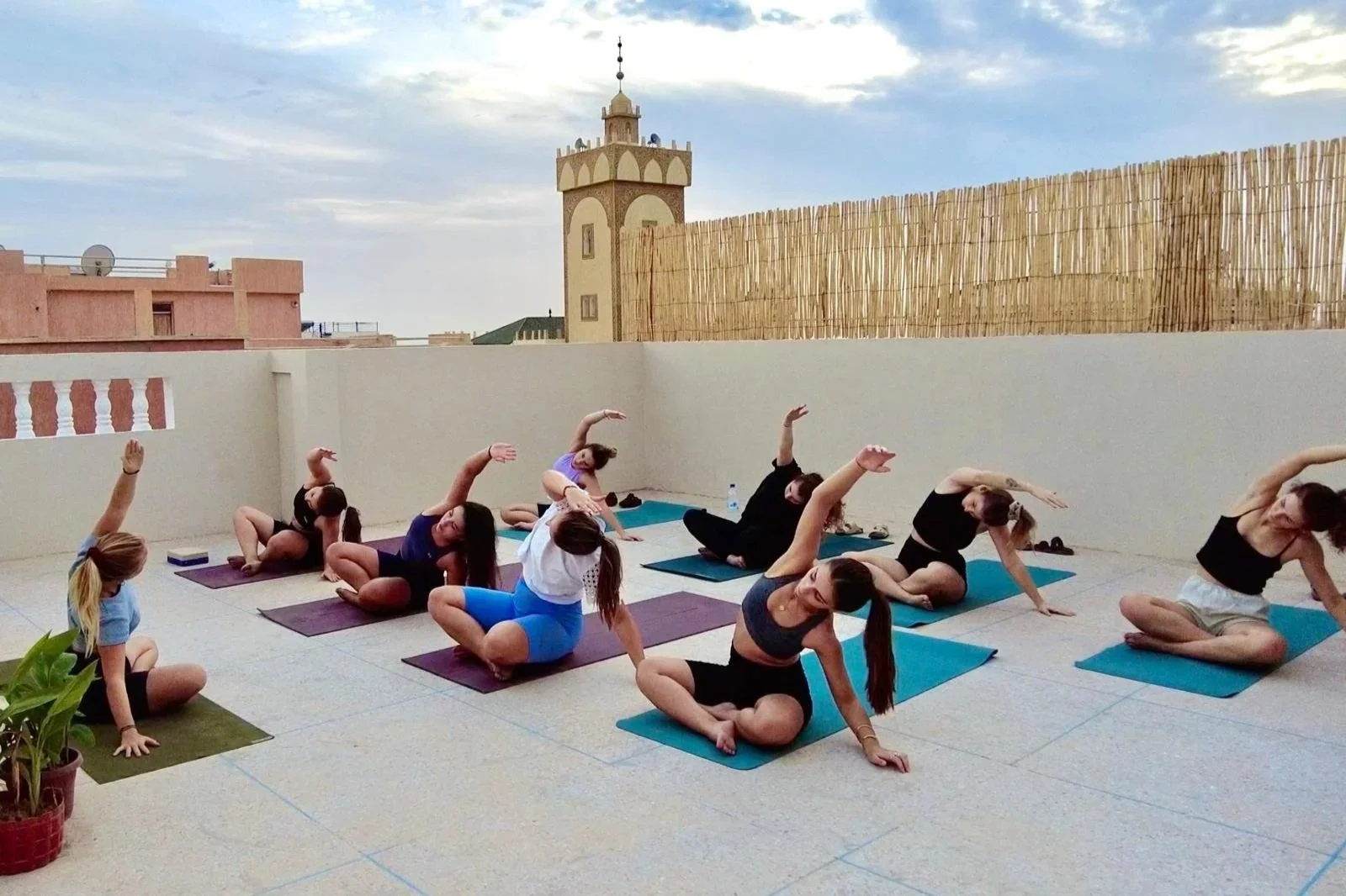 Group of women and girls practicing yoga on a rooftop terrace with cityscape in the background, including a tall tower and cloudy sky.