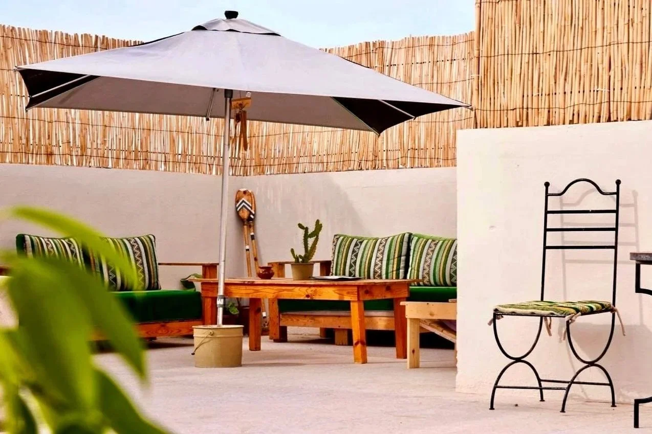 Outdoor patio with striped cushions, a large beach umbrella, a small table with a straw hat, a black metal chair, a wooden table, a cactus, and a bamboo fence in the background.