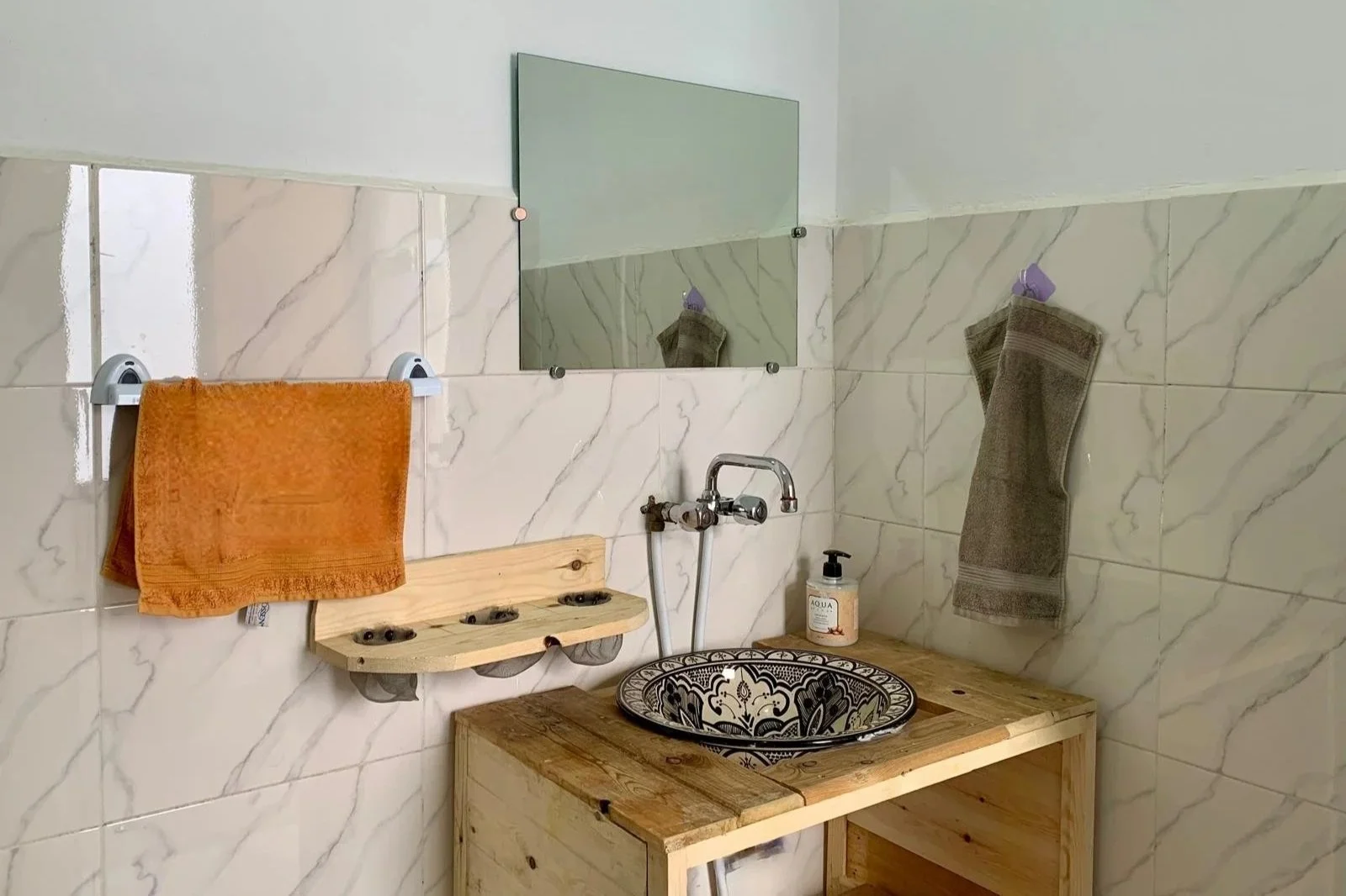 Bathroom with marble walls, a wooden vanity with a decorative bowl sink, towel racks with an orange towel and a gray towel, a small mirror, and a soap dispenser.