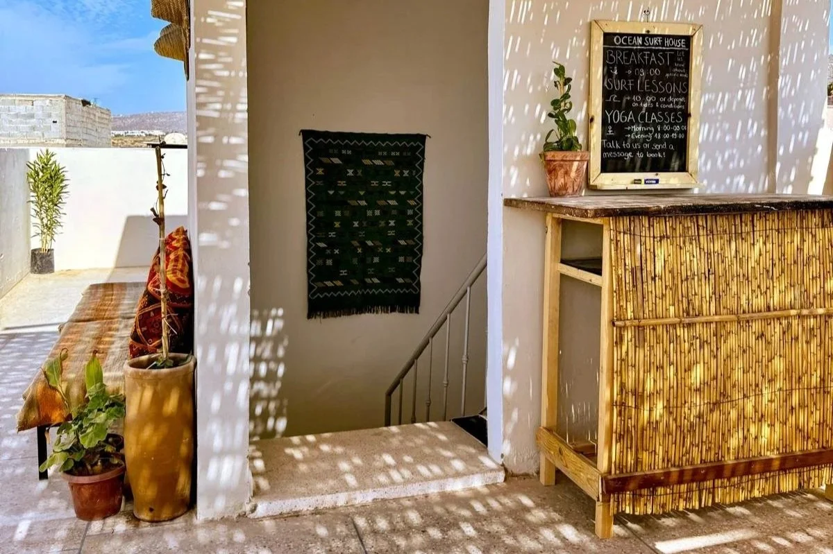 Sunlit outdoor bar area with bamboo decor, potted plants, and a chalkboard menu listing breakfast, surf lessons, and yoga classes at an ocean surf house.