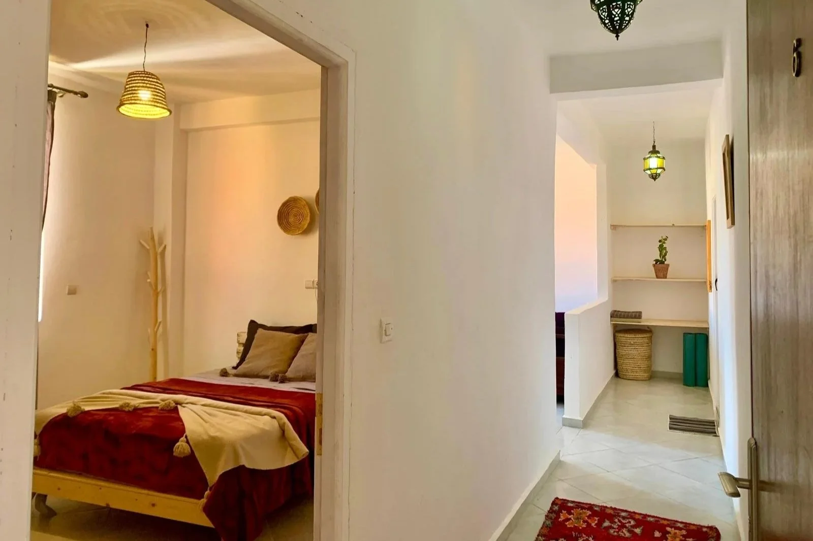 View of a hallway leading to a bedroom in a house or apartment, with decorative hanging lights, white walls, and a red patterned rug on the floor.