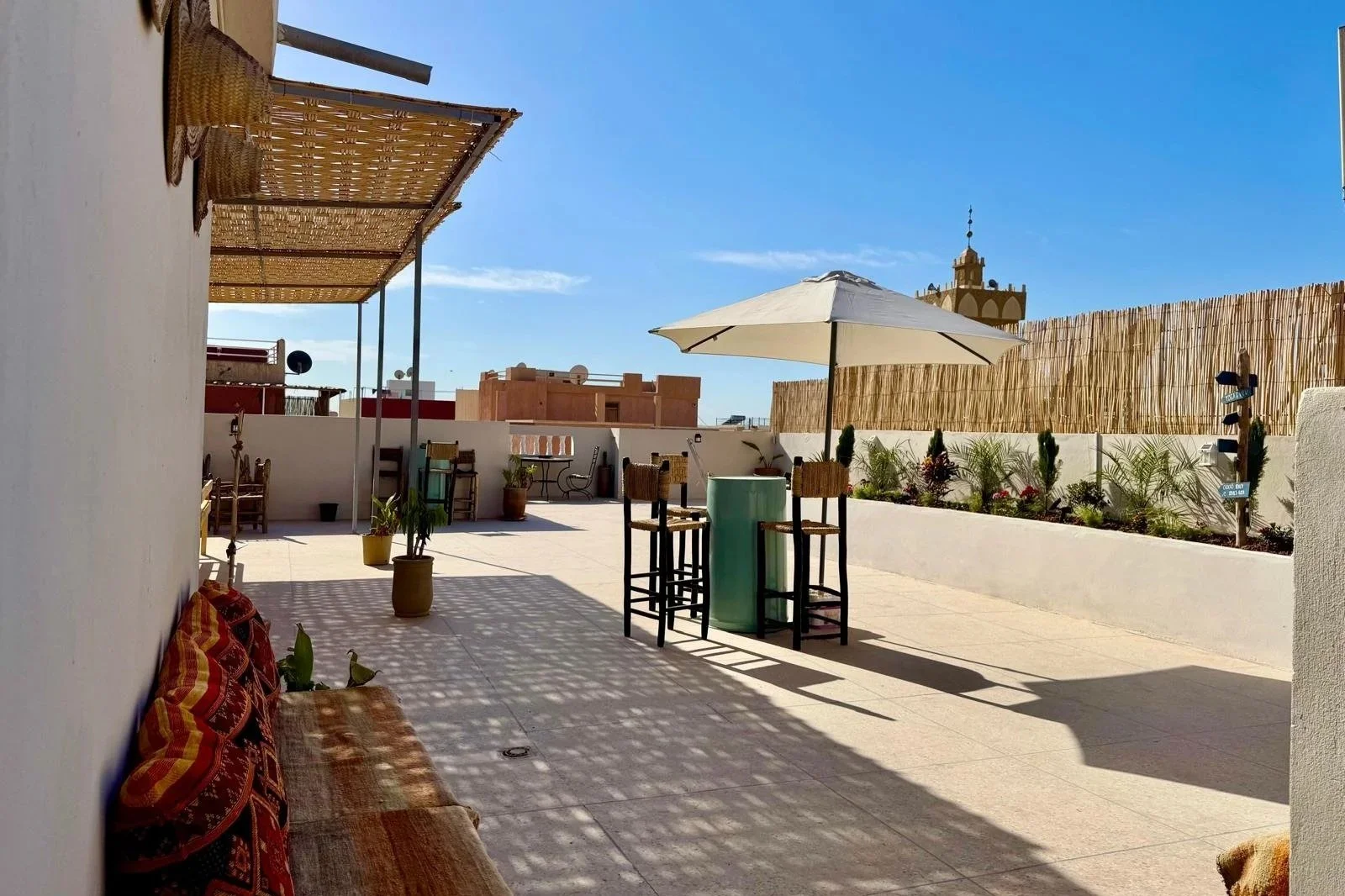 Rooftop patio with some chairs, a table with an umbrella, potted plants, and a decorative wooden wall, under a bright blue sky.