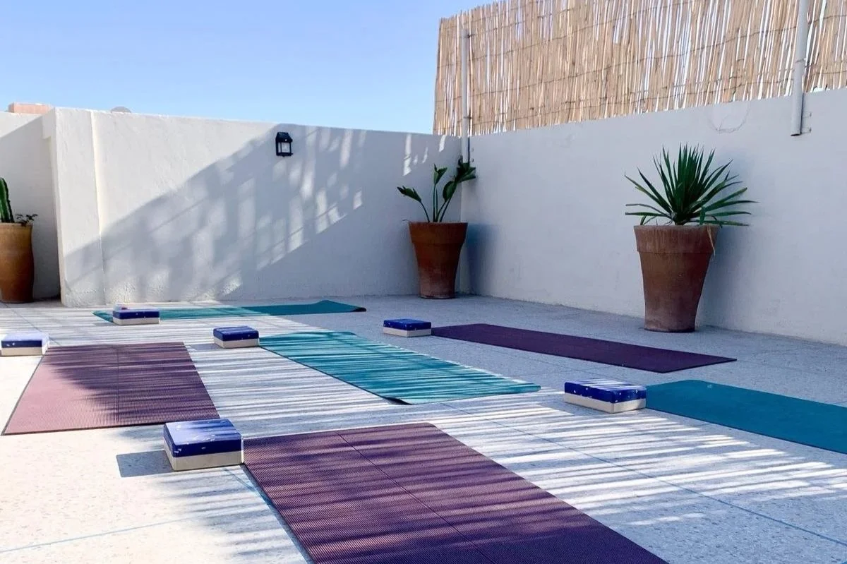 Outdoor yoga setup with yoga mats and blocks on a tiled terrace, potted plants, and a white wall with bamboo fencing and a wall-mounted light.