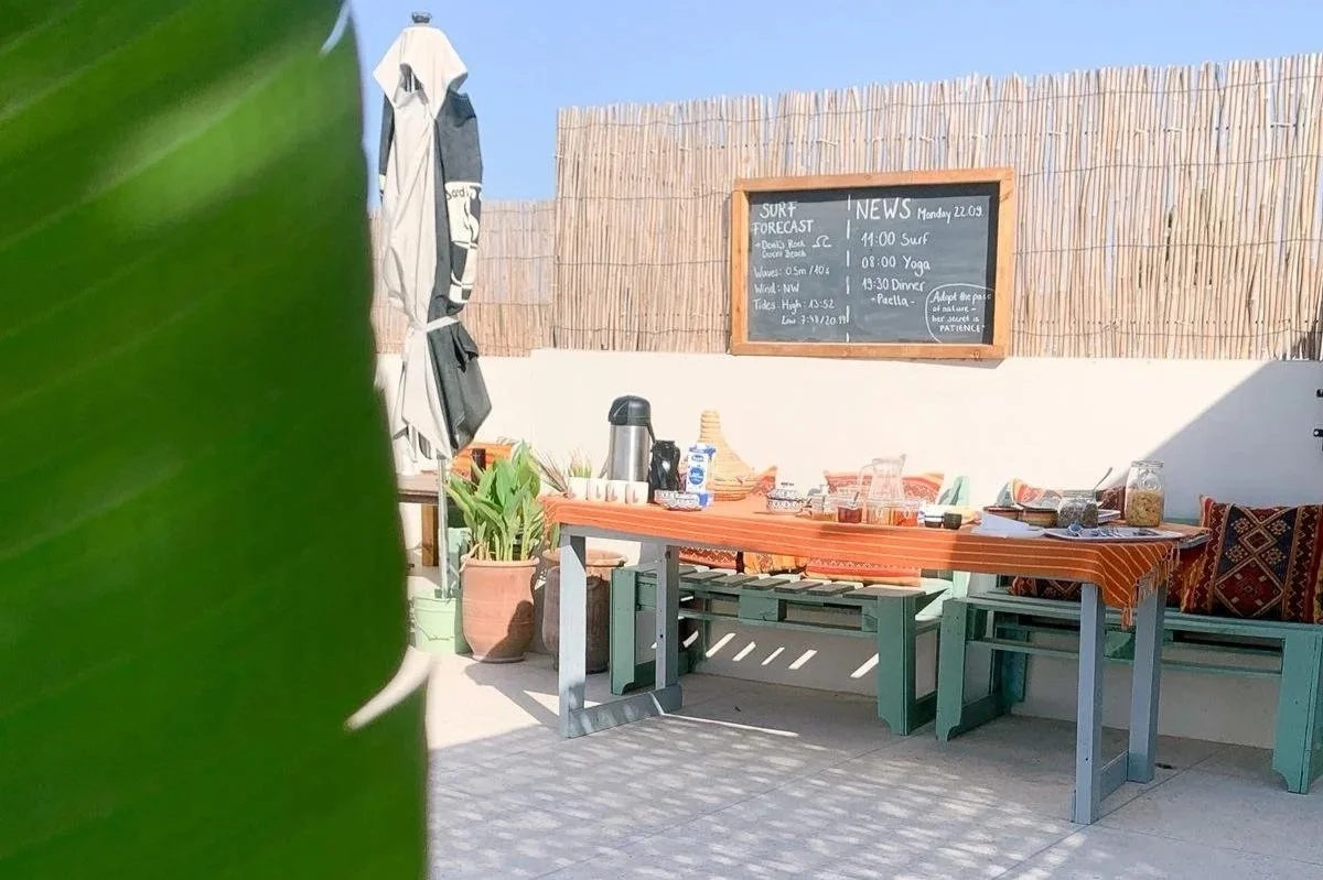 Outdoor seating area with a wooden table, pillows, and various items on the table. There is a blackboard with a surf forecast and news, and a large green leaf partially obstructs the view in the foreground.
