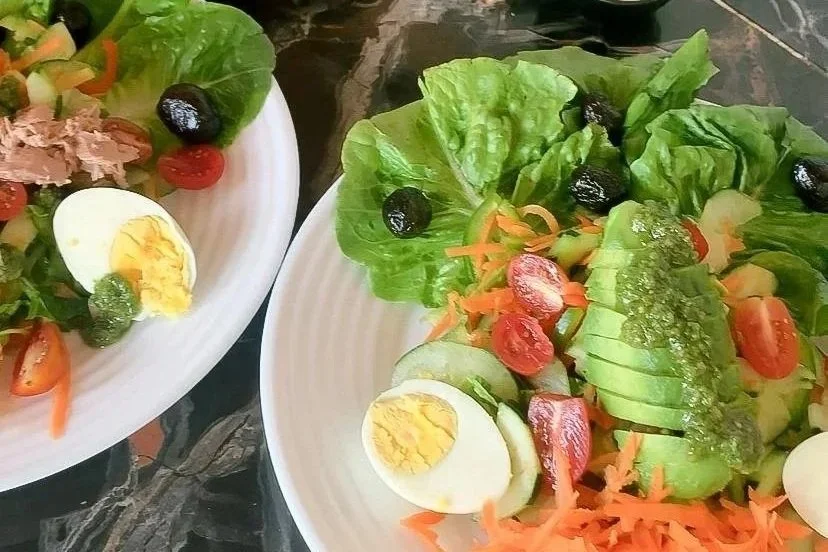 Two plates of fresh vegetable salads with lettuce, cherry tomatoes, sliced cucumbers, shredded carrots, black olives, half boiled eggs, and green dressing.