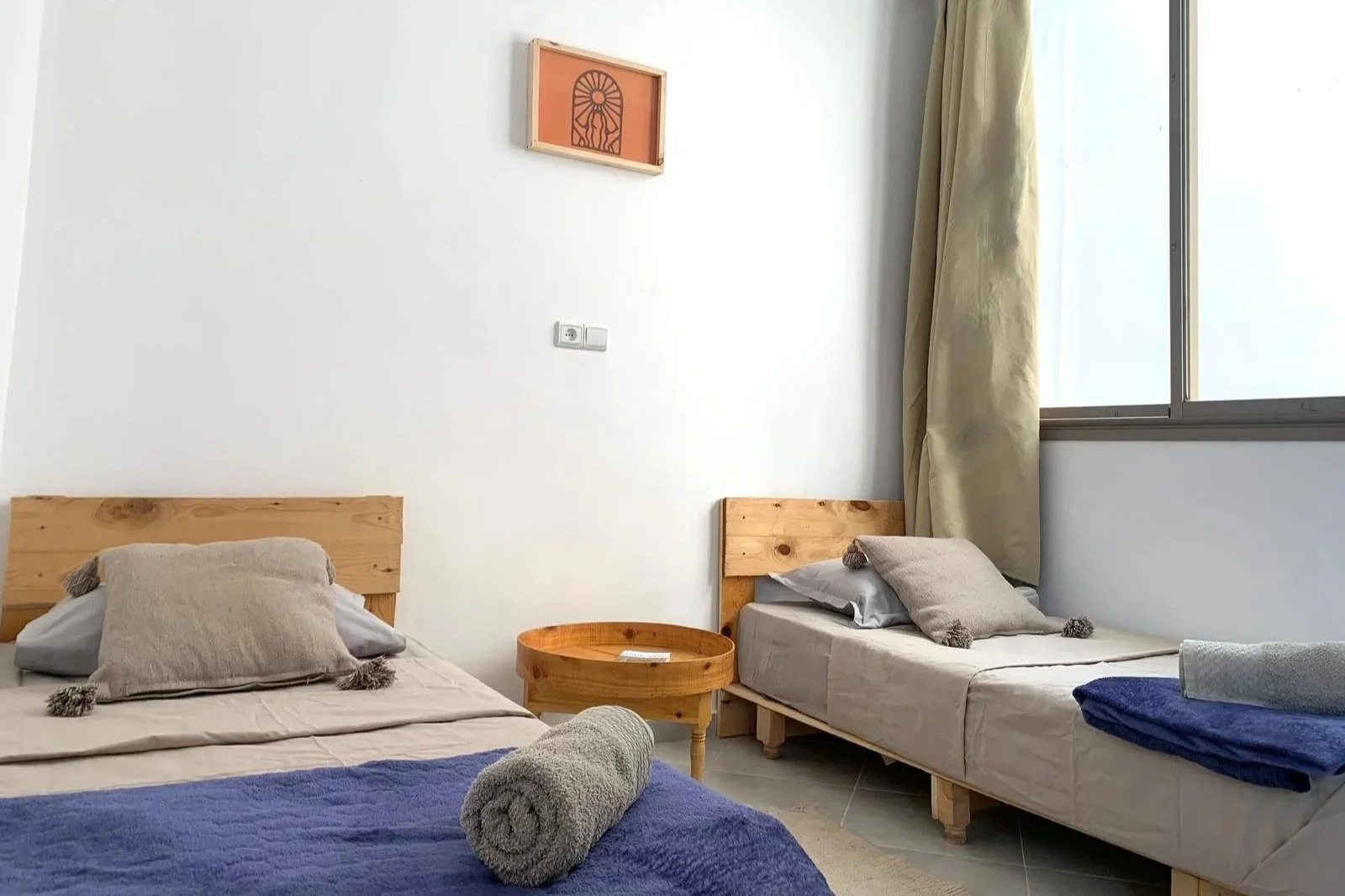 A bedroom with two single beds, beige bedding, and gray pillows with tassels. A rolled gray towel is on the bed in the foreground, covered partially by a blue blanket. A round wooden nightstand is between the beds. A window with beige curtains is on 