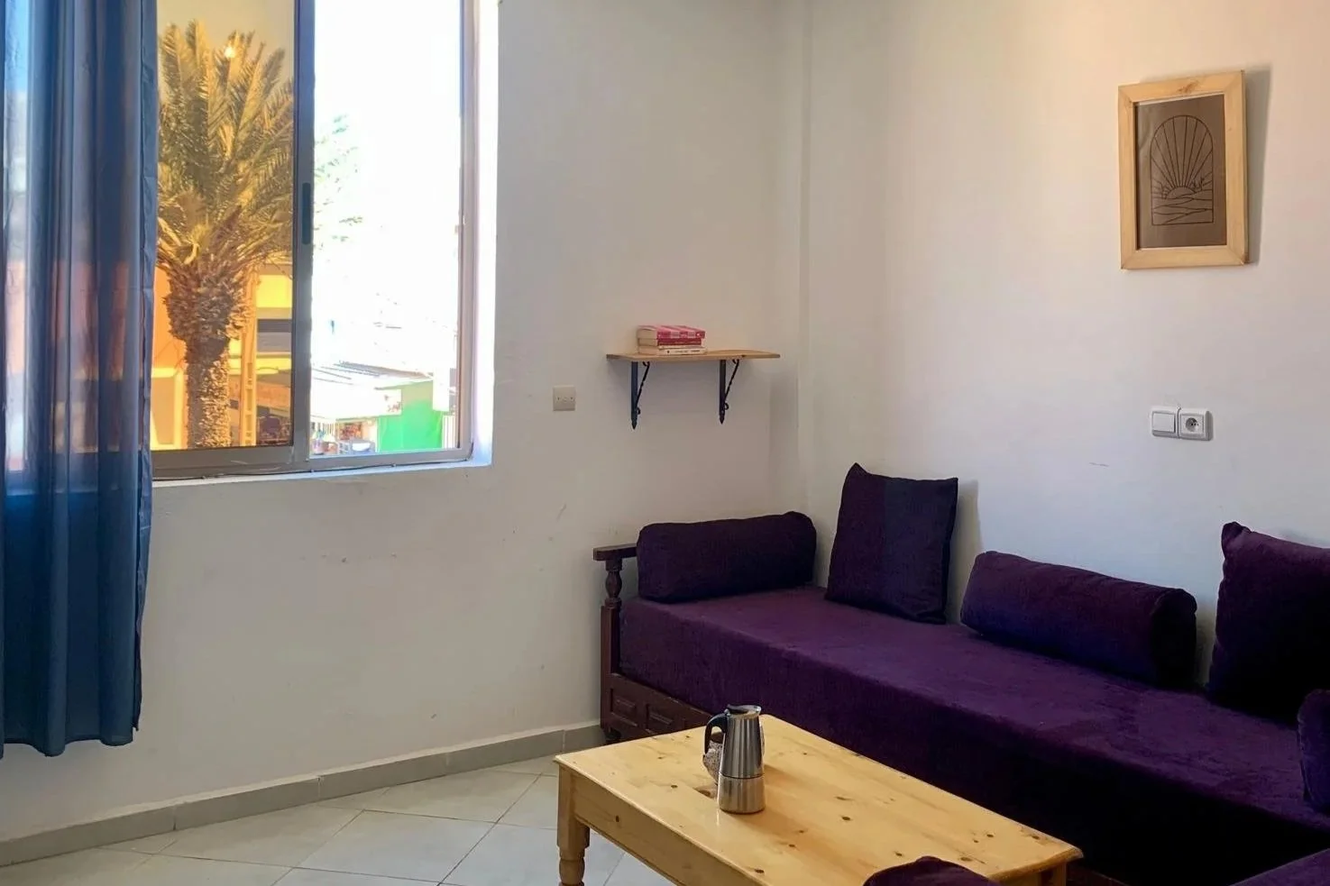 Living room with a window, purple sofas, a wooden coffee table with a coffee pot, a framed picture on white wall, a small shelf with books, and a rattan ceiling light.