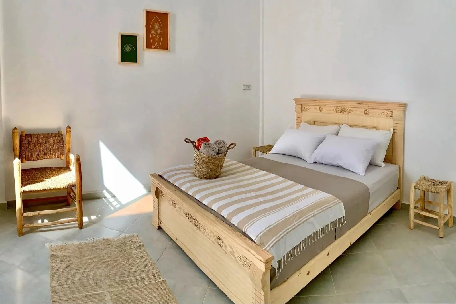 A bedroom with a wooden bed, white pillows, a basket of rolled towels on the bed, a small wooden chair, and wall art, with sunlight casting a glow on the floor.