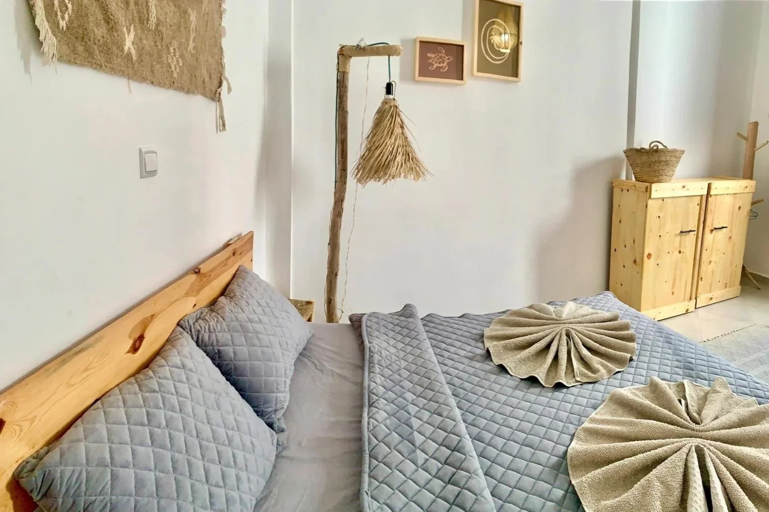 A neatly made bed with gray quilted bedding and pillows in a cozy room decorated with light-colored wooden furniture, wall art, and rustic decor elements.