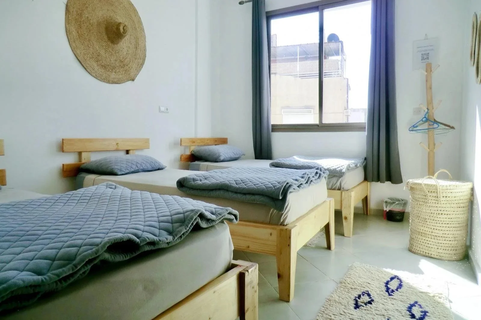 A bedroom with four single beds, each with a wooden frame, gray quilted blankets, and pillows. A large window with blue curtains lets in natural light. A round woven wall hanging, a wooden coat rack, a woven basket, and a small trash bin are visible 