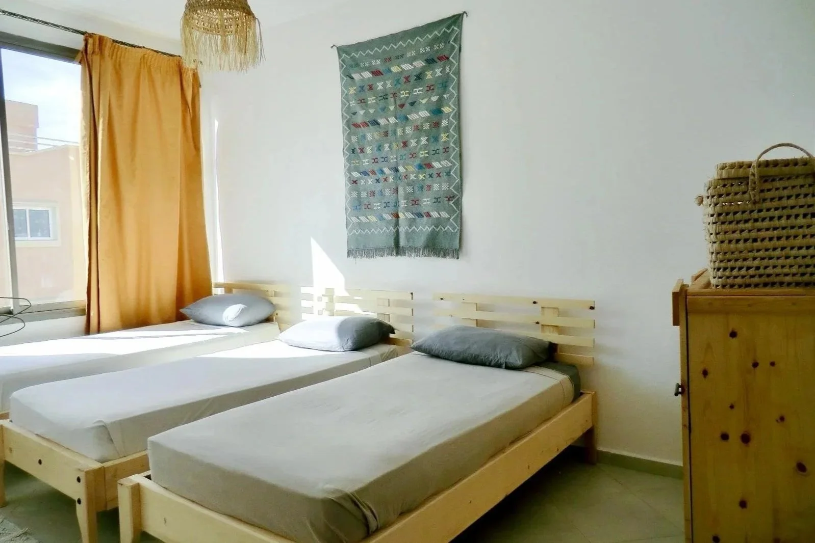 A bedroom with three single beds, gray pillows, a window with yellow curtains, a woven light fixture, a woven wall hanging, and a wooden dresser with a wicker basket on top.