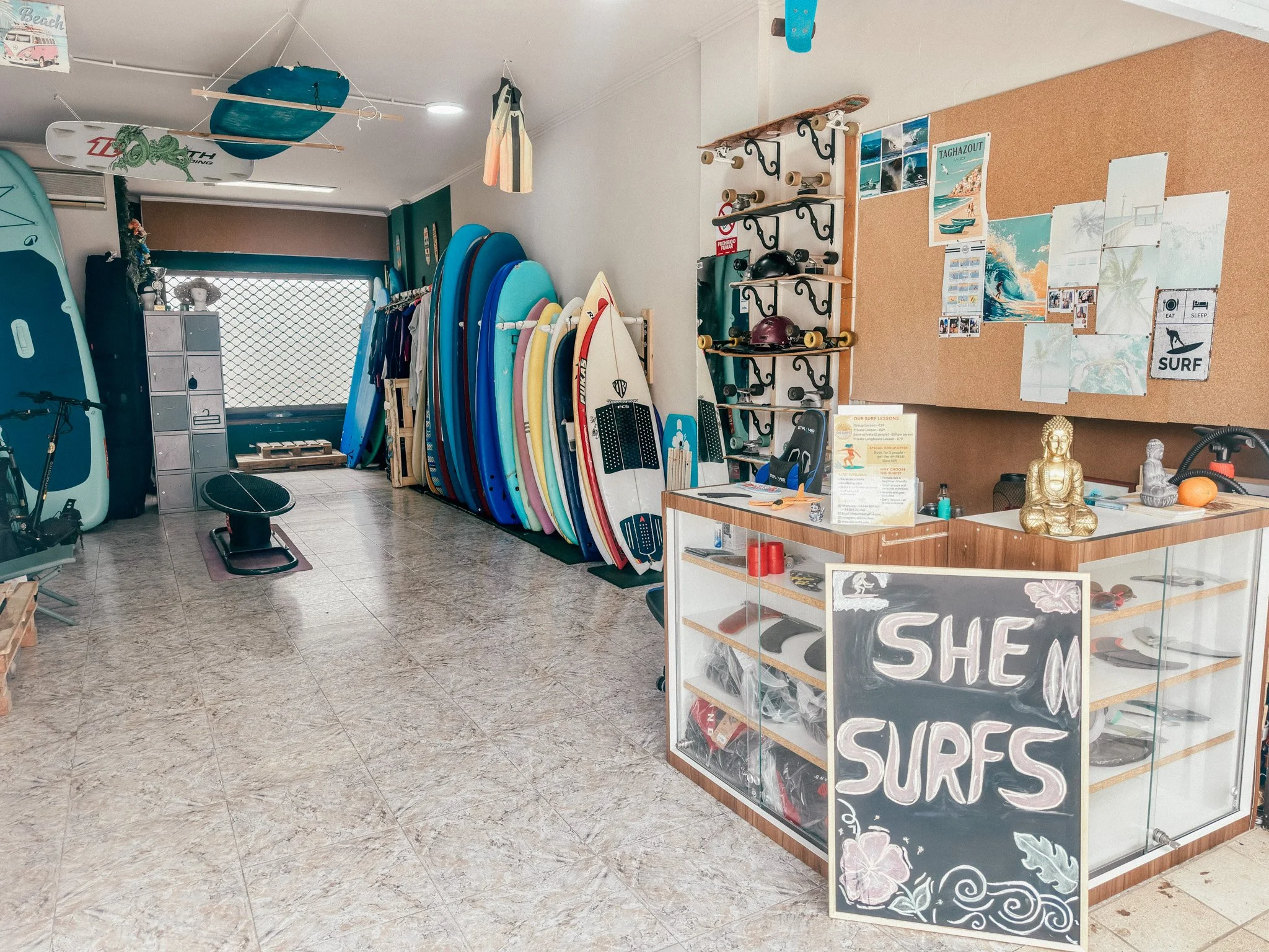 Indoor surf shop with surfboards on display, a chalkboard sign that reads 'SHE N SURFS', lockers, and surfing accessories on shelves.