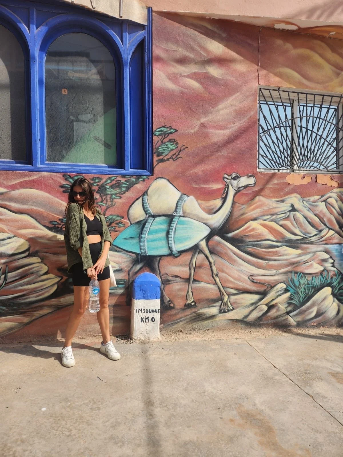 A woman with sunglasses, holding a water bottle, standing in front of a colorful mural of a camel with a large blue shell on its back and a desert landscape.