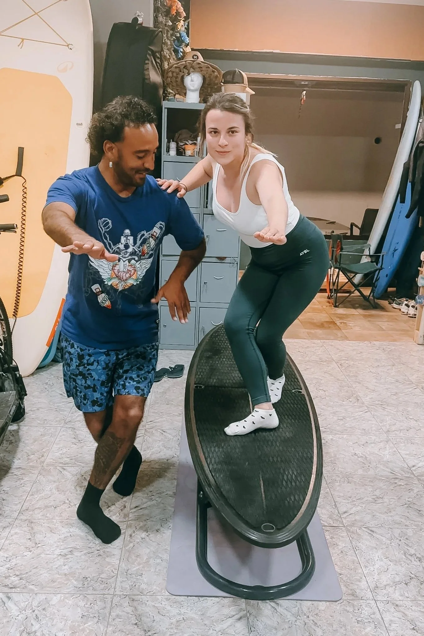 A woman balancing on a balance board with her arms extended, being assisted by a man who is guiding her. The scene takes place indoors, with fitness equipment and surfboards in the background.