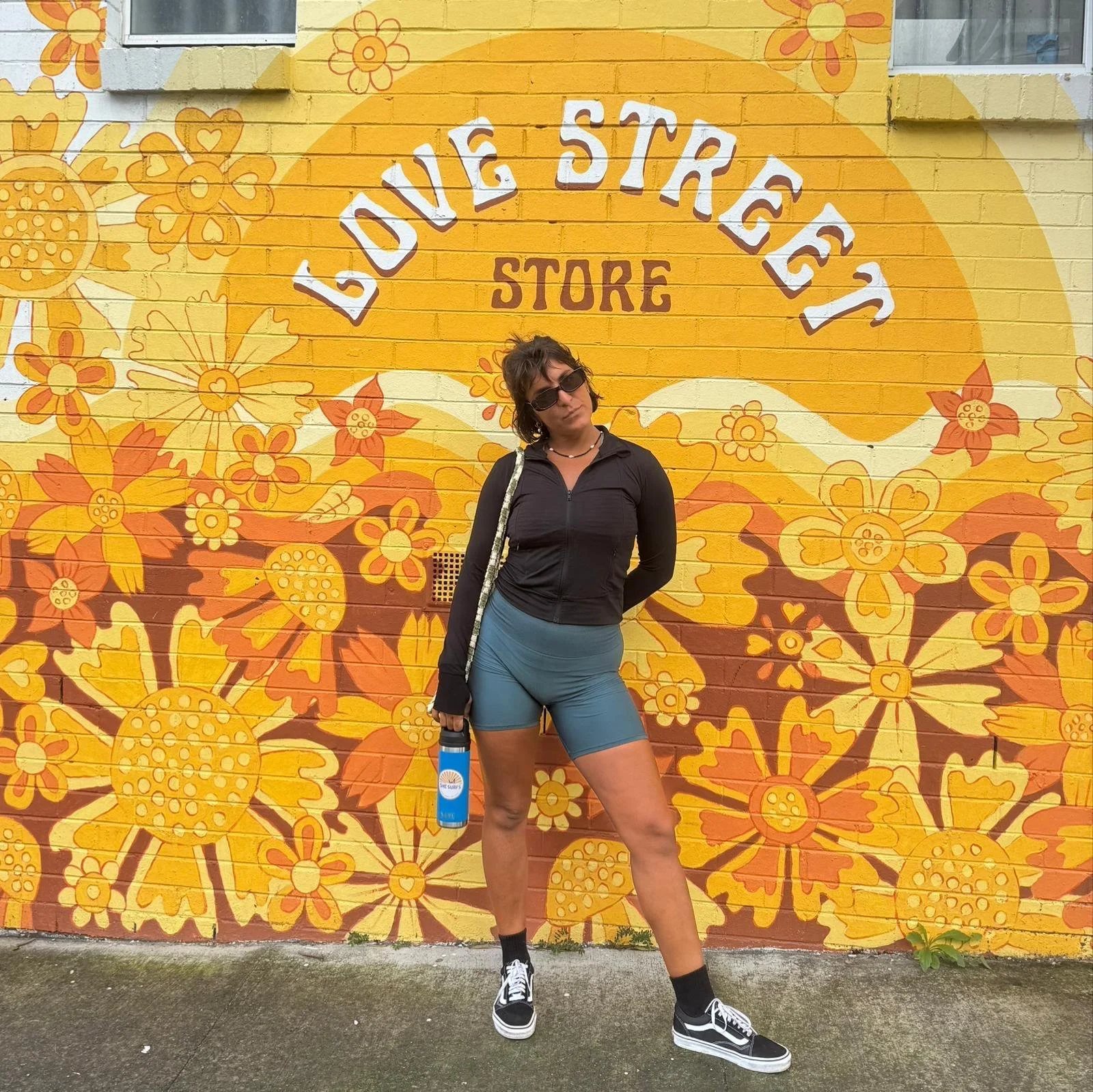 A woman in athletic wear standing in front of a brightly painted yellow and orange floral mural with the words 'Love Street Store' on it, holding a blue water bottle and wearing sunglasses.