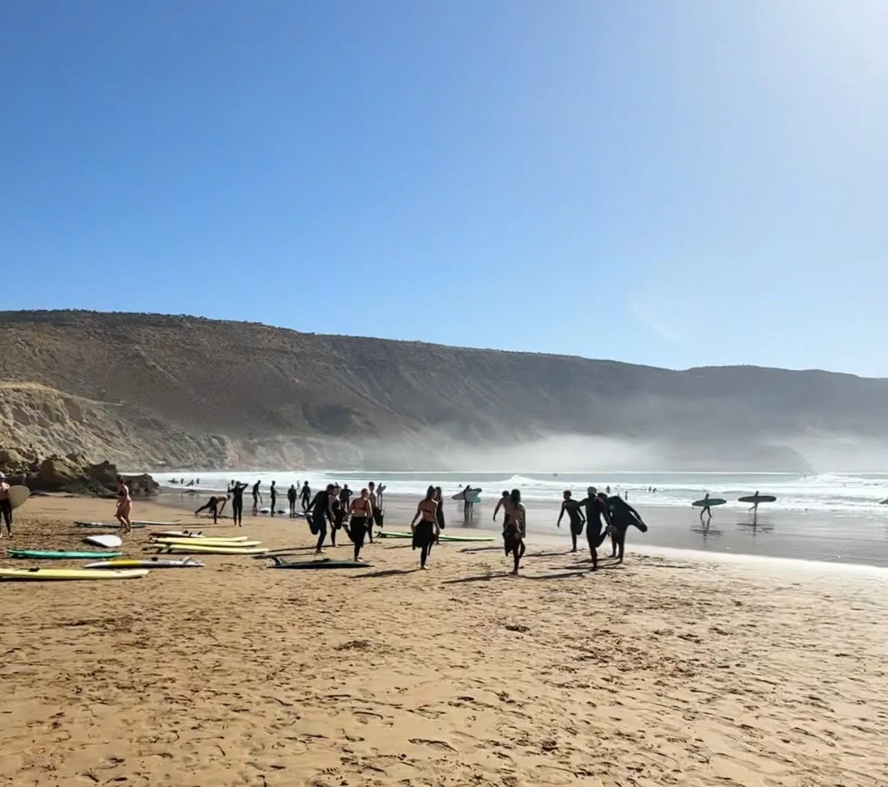 People carrying surfboards walking on a sandy beach with more surfers in the water, cliffs in the background, and a clear blue sky.