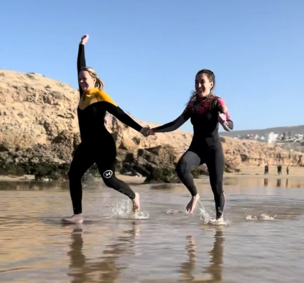 Two women in wetsuits holding hands and running through shallow water on a beach, with cliffs and a clear blue sky in the background.