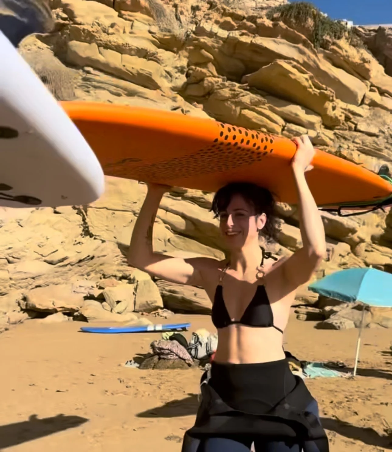 A woman in a black bikini holding an orange surfboard above her head on a sandy beach with rocks behind her.
