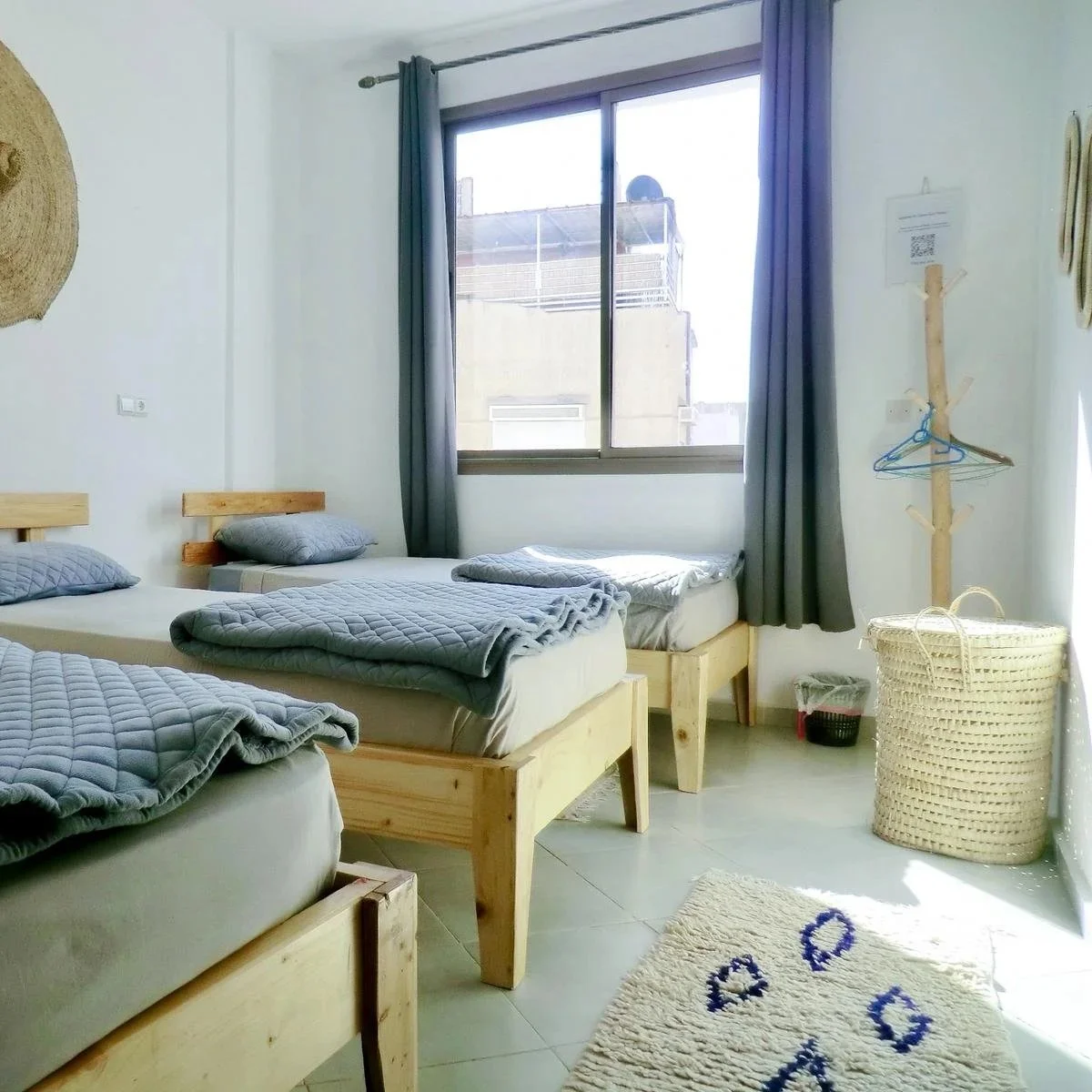 A bedroom with three single beds made of light wood, each with gray quilted blankets and pillows. There's a large window with gray curtains, a wicker laundry basket, a wooden coat stand with hangers, and a cream-colored rug on the tiled floor.
