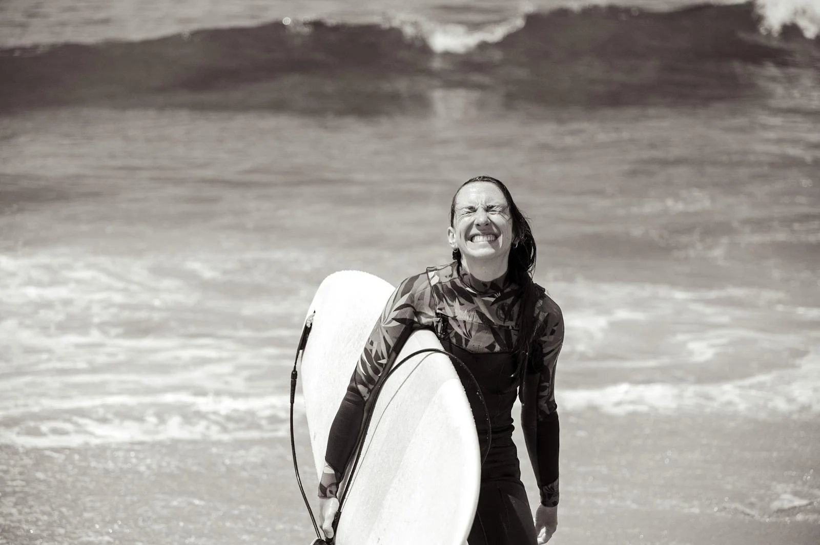 Woman smiling with a surfboard on a beach, wearing a wetsuit, and standing in front of the ocean waves.