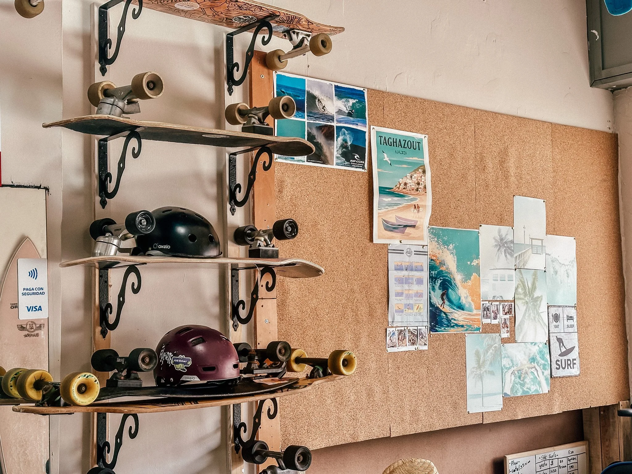 Wall display of skateboards with helmets on a rack, and a corkboard featuring surfing-themed posters and photos.