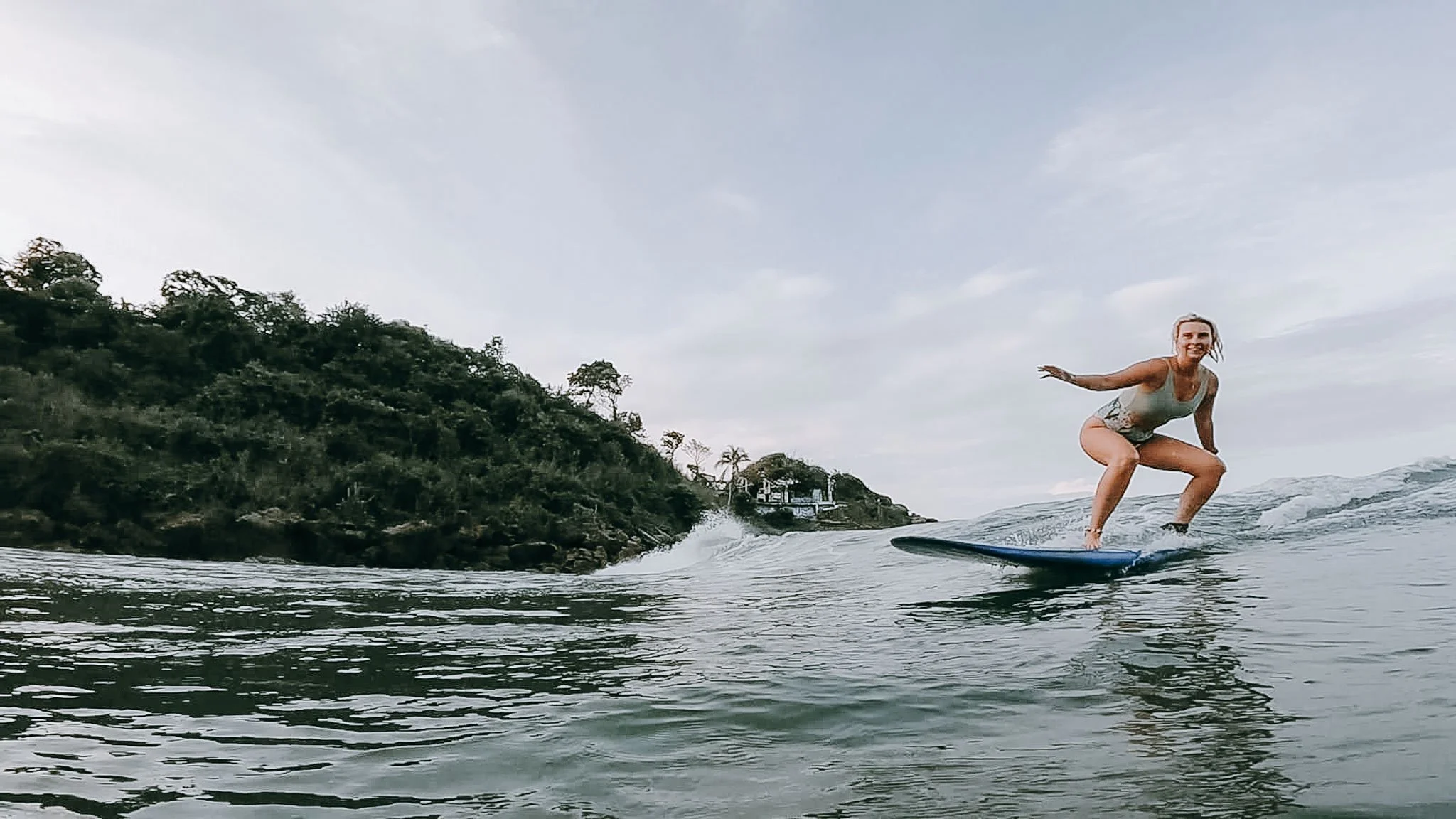 Woman surfing on a wave near a forested coastline during daytime.