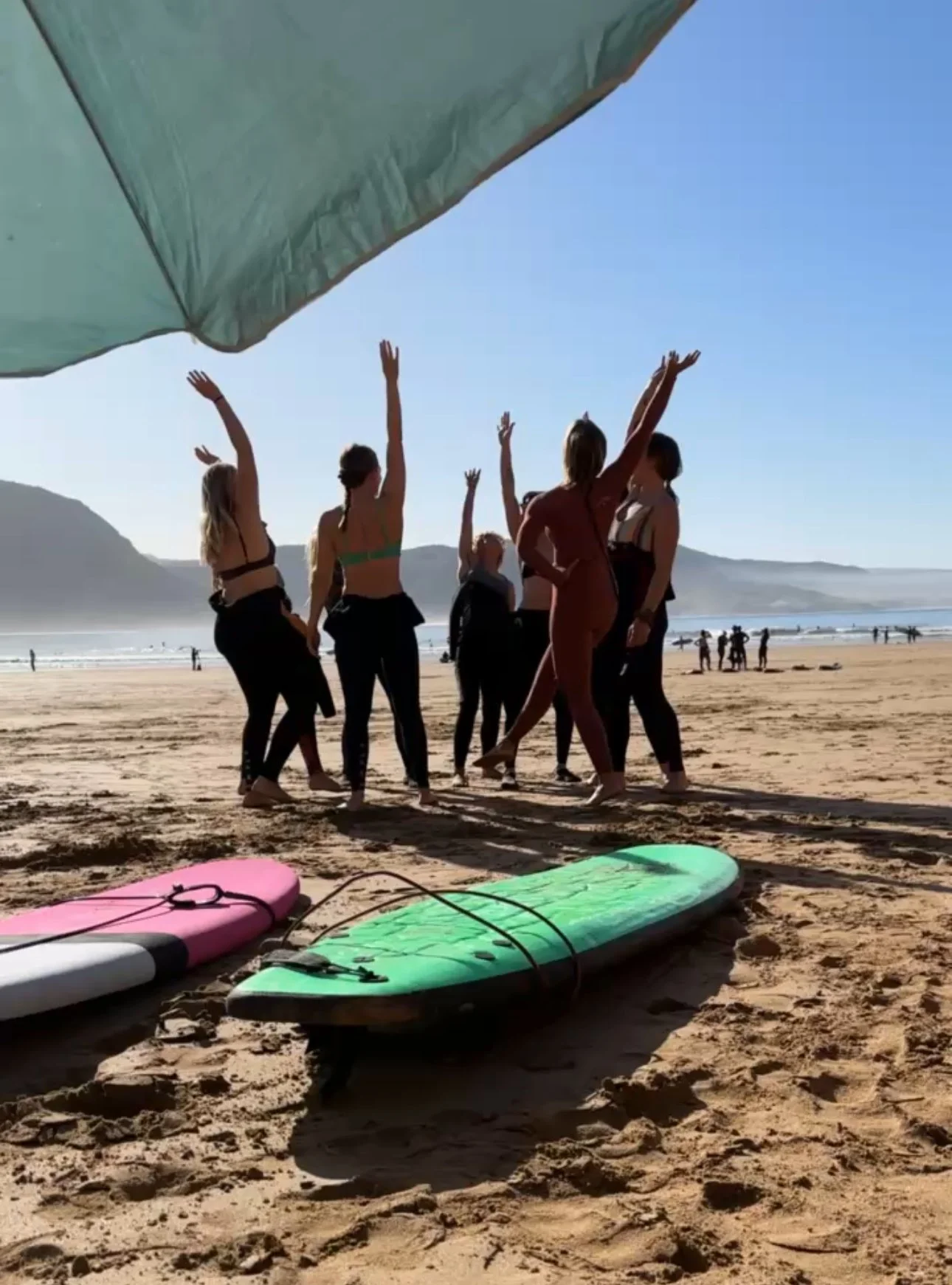 Group of people dancing on the beach under a large beach umbrella, with surfboards on the sand and mountains in the background.