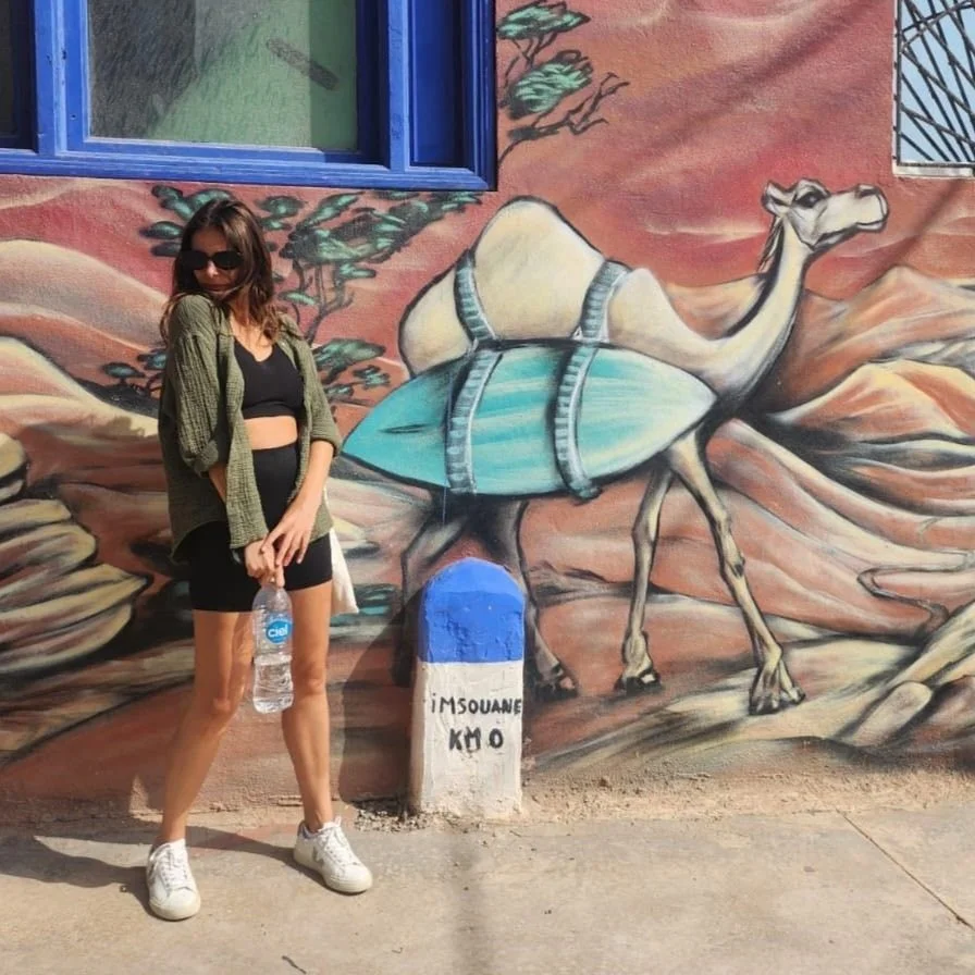 Young woman in black sports bra, black shorts, sunglasses, and green jacket standing next to a colorful mural of a camel painted on a wall.