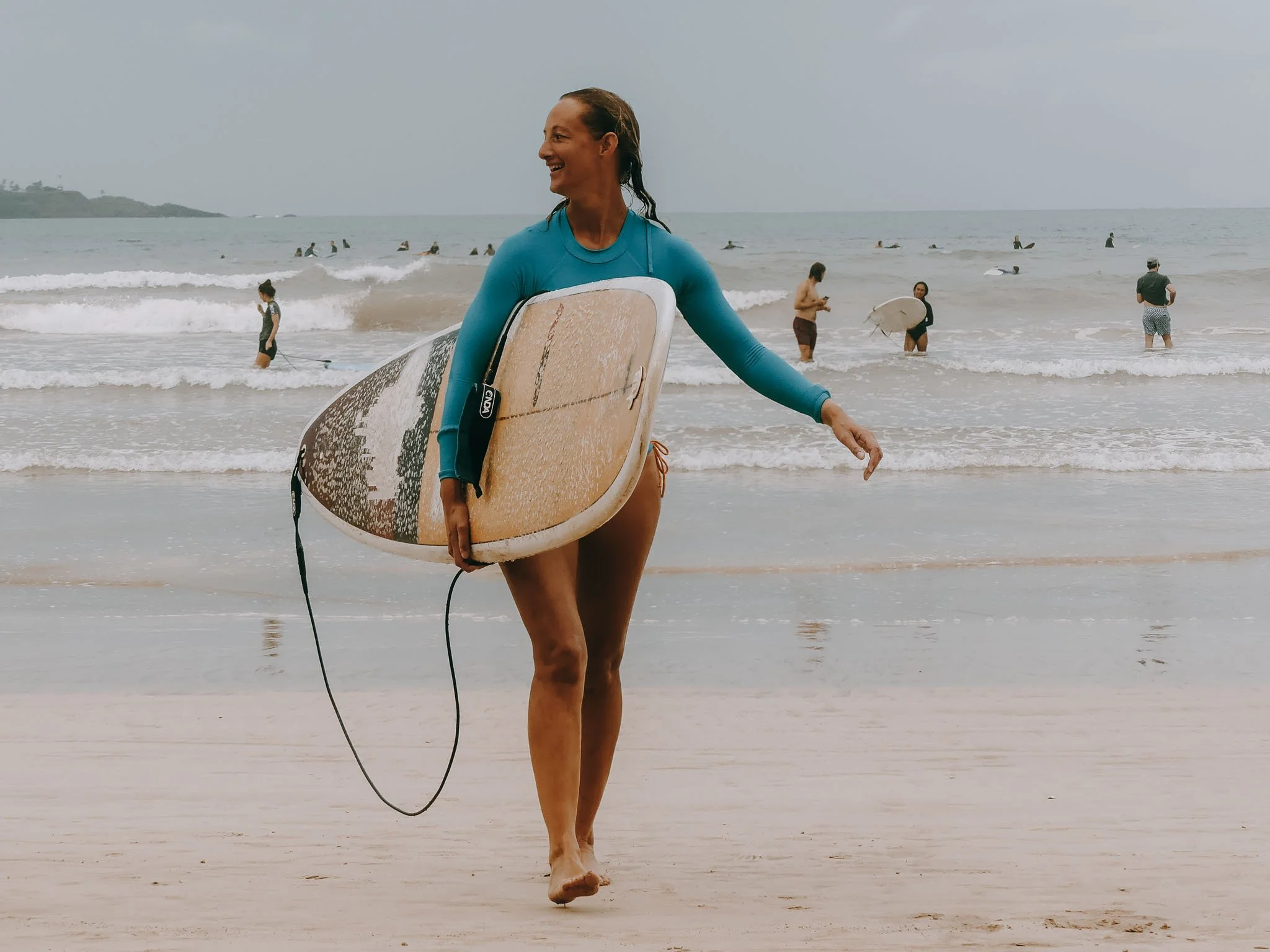 A woman in a blue long-sleeve shirt walking on the beach, carrying a surfboard.