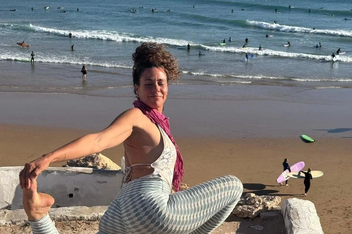 A woman practicing yoga outdoors on a beach with surfers in the ocean in the background.