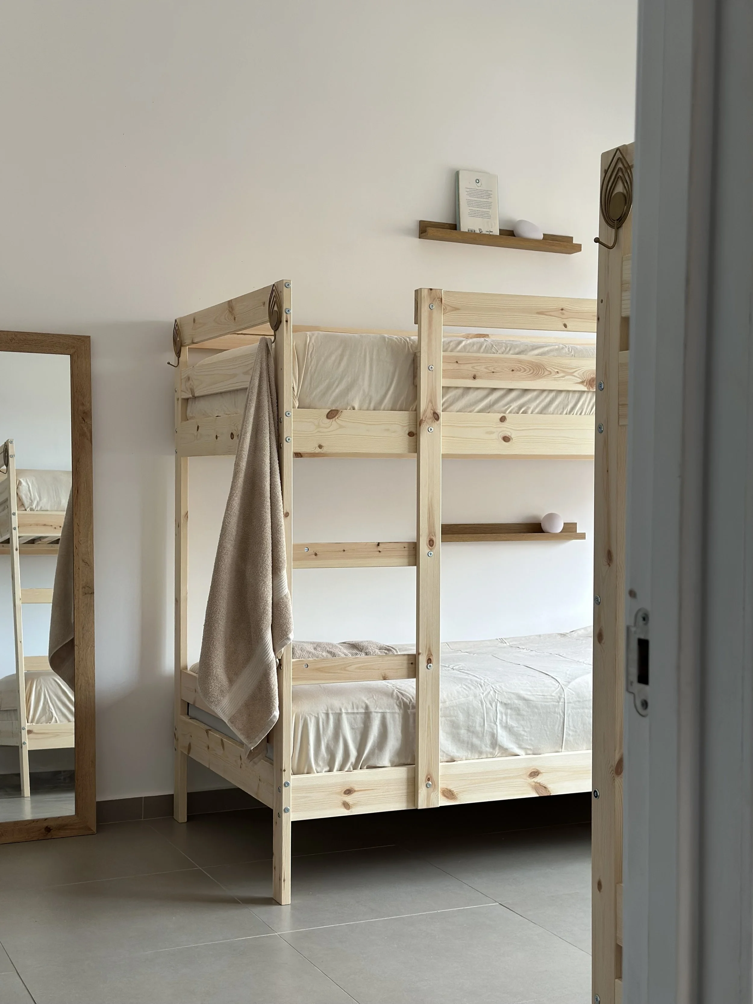 Wooden bunk bed with beige bedding, towel hanging on side, and a mirror on the wall next to it, in a minimalist room with a floating shelf and an abstract decor piece.