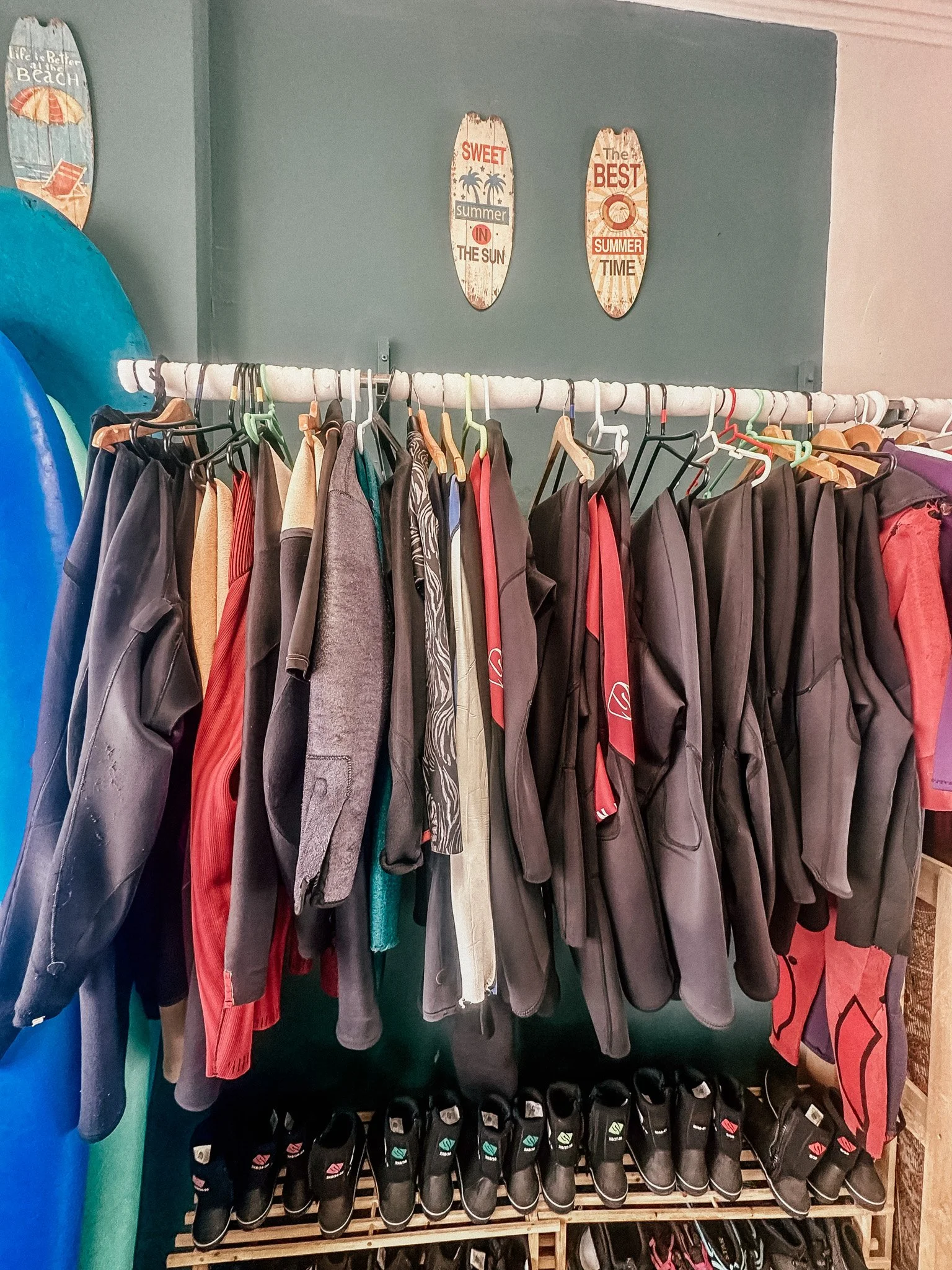 Clothing rack with jackets, hoodies, and shirts, with black flip-flops and shoes on the floor underneath, near a wall with beach-themed signs and surfboards.