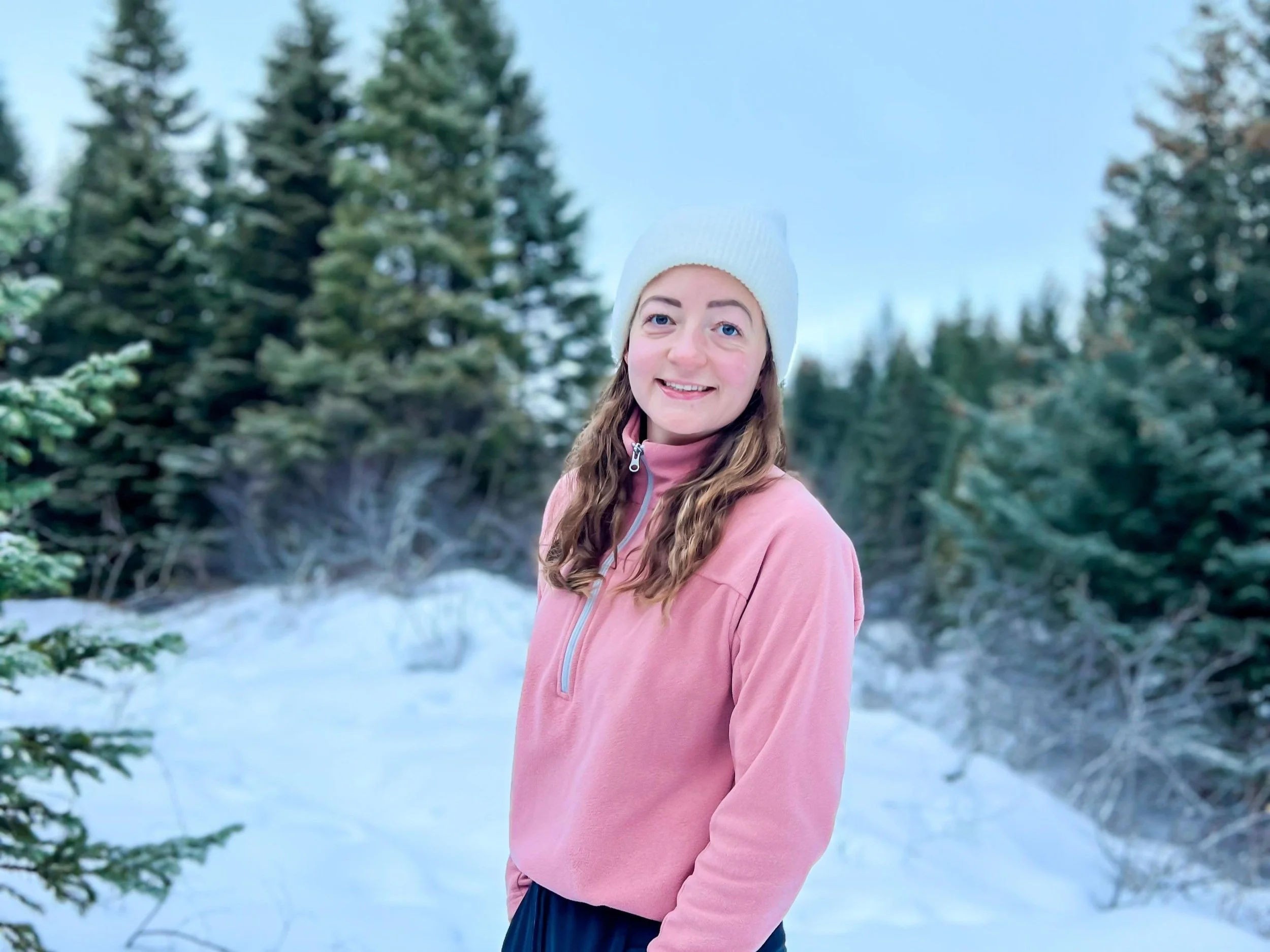 Young woman smiling outdoors in winter, wearing a pink jacket and white knit hat, surrounded by snow and evergreen trees.