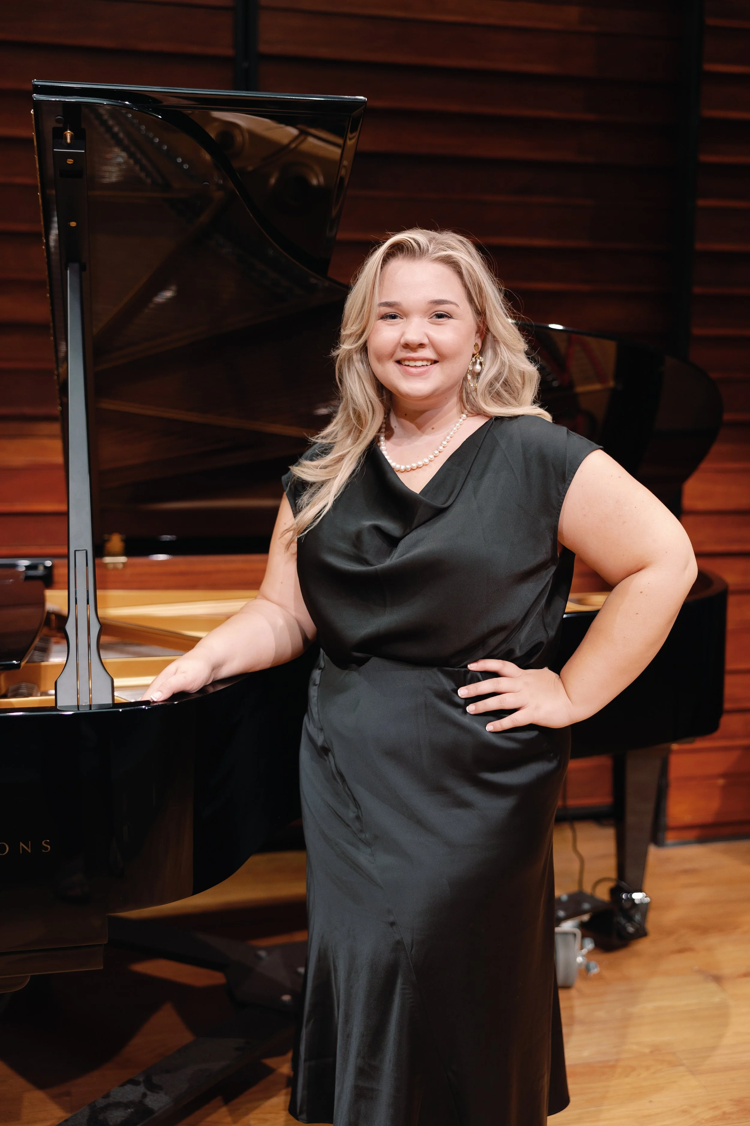 A woman in an elegant black dress stands next to a grand piano in a wood-paneled room.