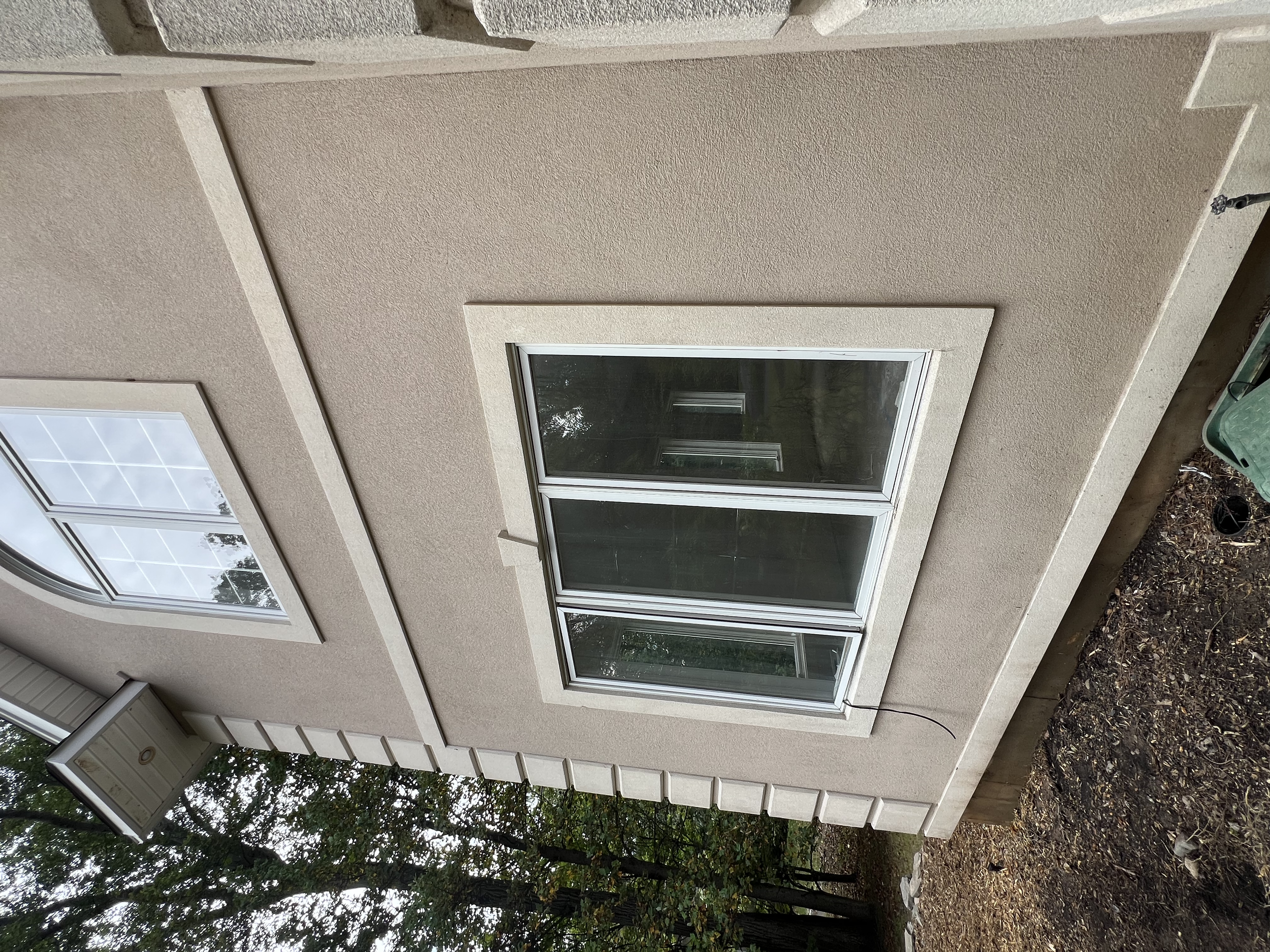 A beige stucco house exterior with a large rectangular window and a smaller arched window, surrounded by trees and a patch of dirt ground. See the difference professional house washing makes! Our soft wash system removes dirt, mold, algae, and mildew