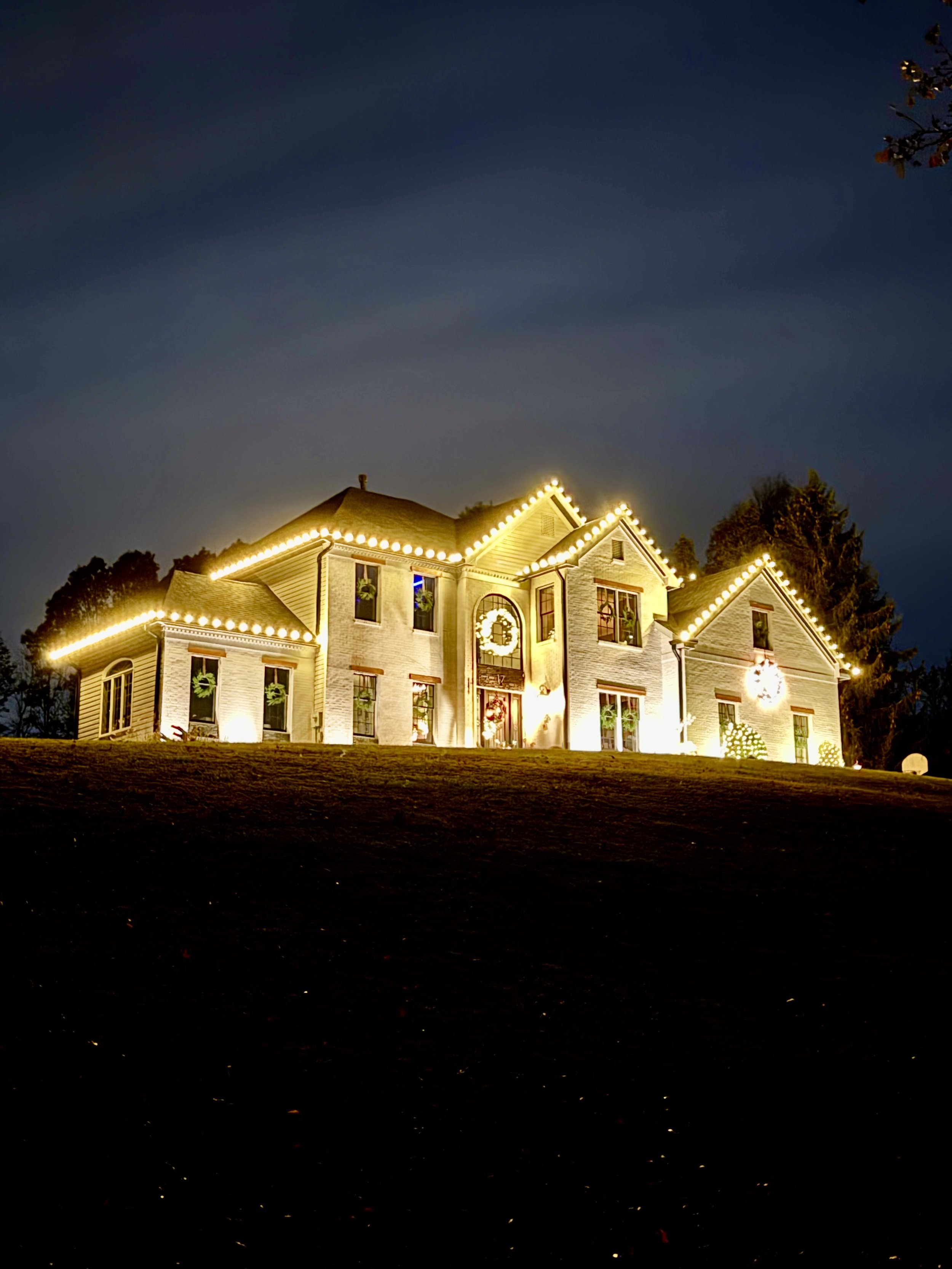 Large house decorated with Christmas lights and wreaths at night. A house decorated with Christmas lights at night, including lights outlining the roof and garage, a illuminated wreath on the front, and decorated Christmas trees outside. Professional