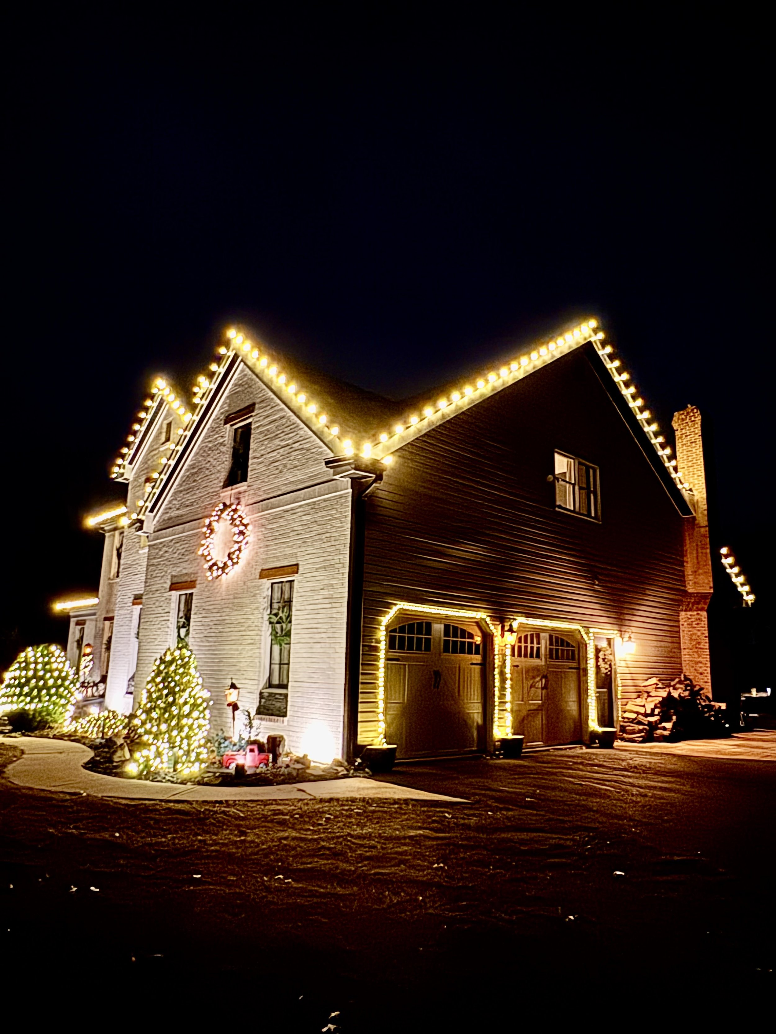 A house decorated with Christmas lights at night, including lights outlining the roof and garage, a illuminated wreath on the front, and decorated Christmas trees outside. Professional Christmas lights installation service. 