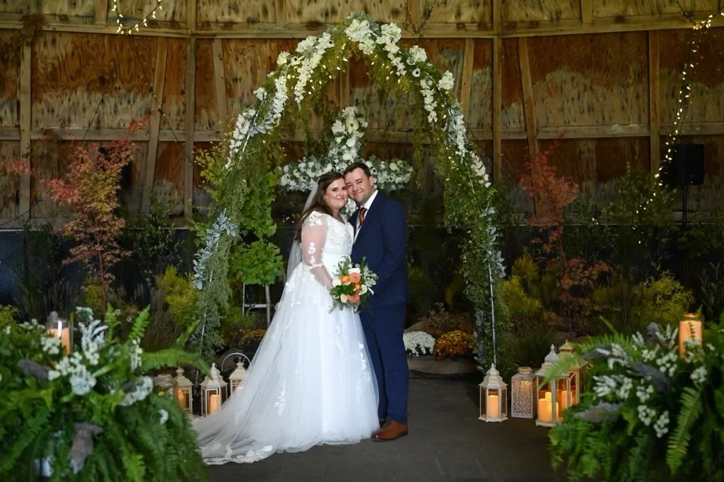 A bride and groom standing under a floral arch during wedding ceremony in a rustic barn, surrounded by greenery and candles.