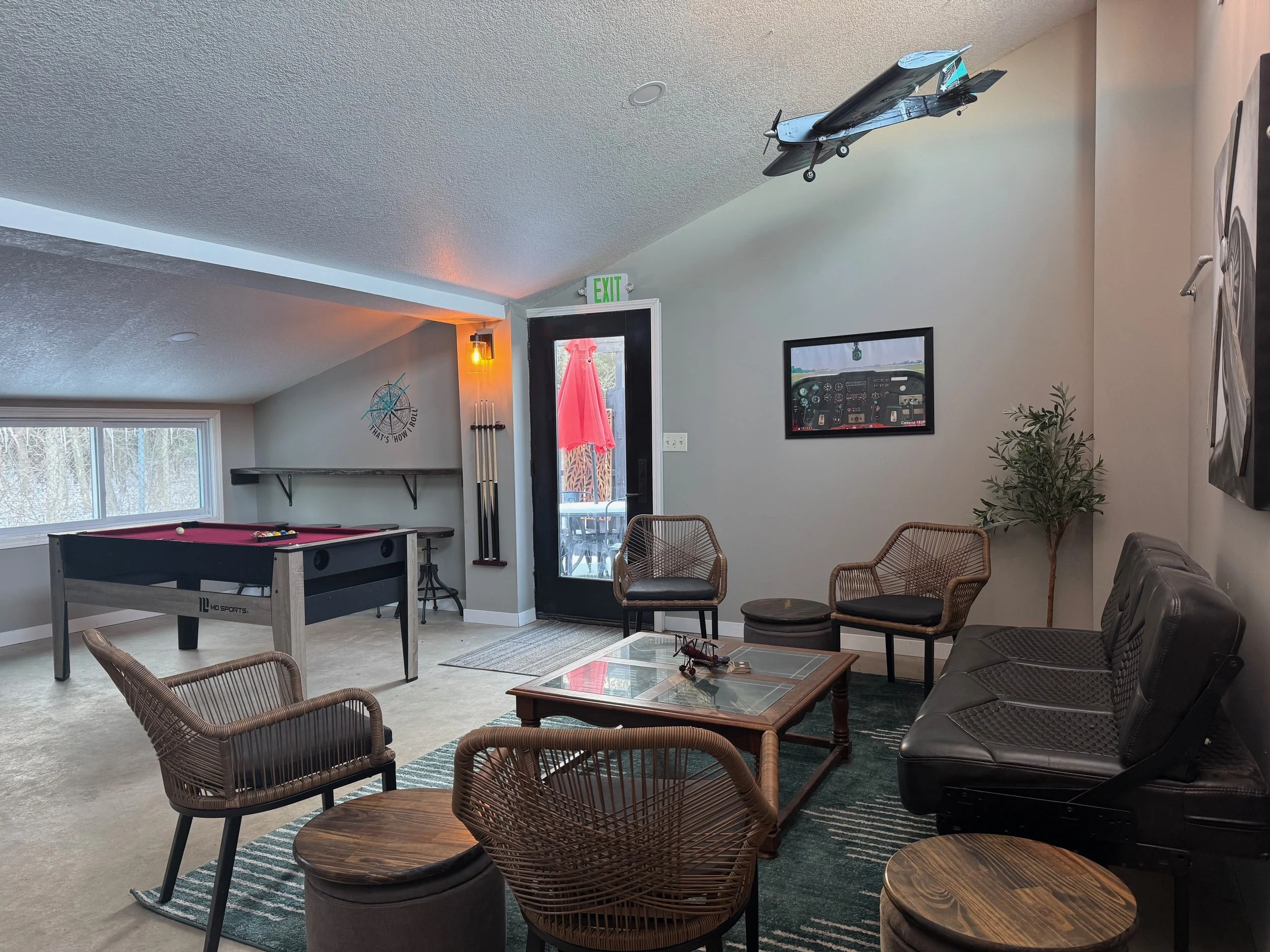 Interior of a recreational room with a pool table, wicker chairs, a couch, and wall decorations including a framed picture of an airplane cockpit and a mirror. There is a door with a pink dress hanging outside and an orange wall-mounted light. A model airplane is mounted on the ceiling.