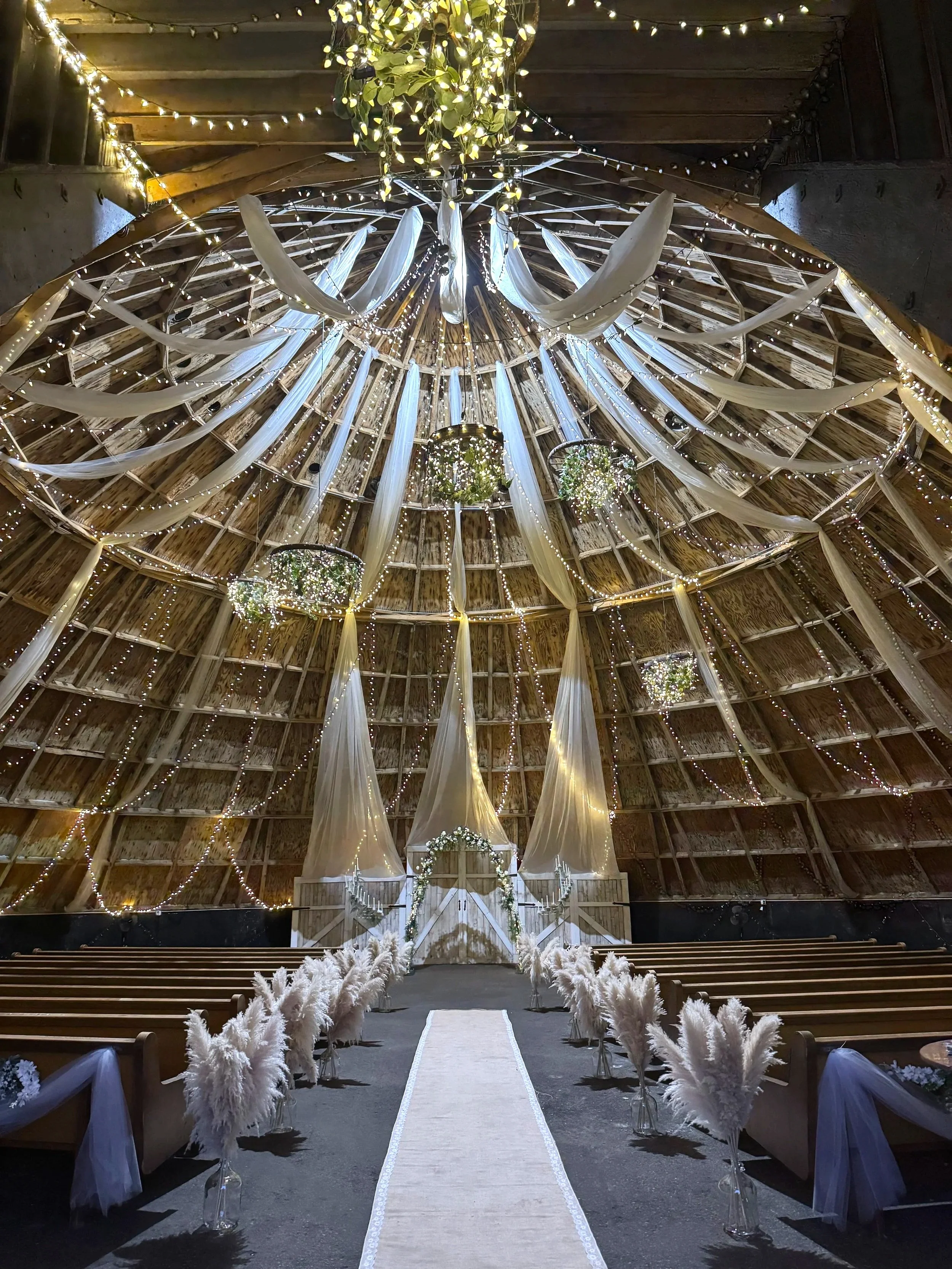 A decorated wedding venue inside a barn with a high wooden ceiling, fairy lights, white drapes, floral arrangements, a central aisle with a white runner, and rows of seating on either side.