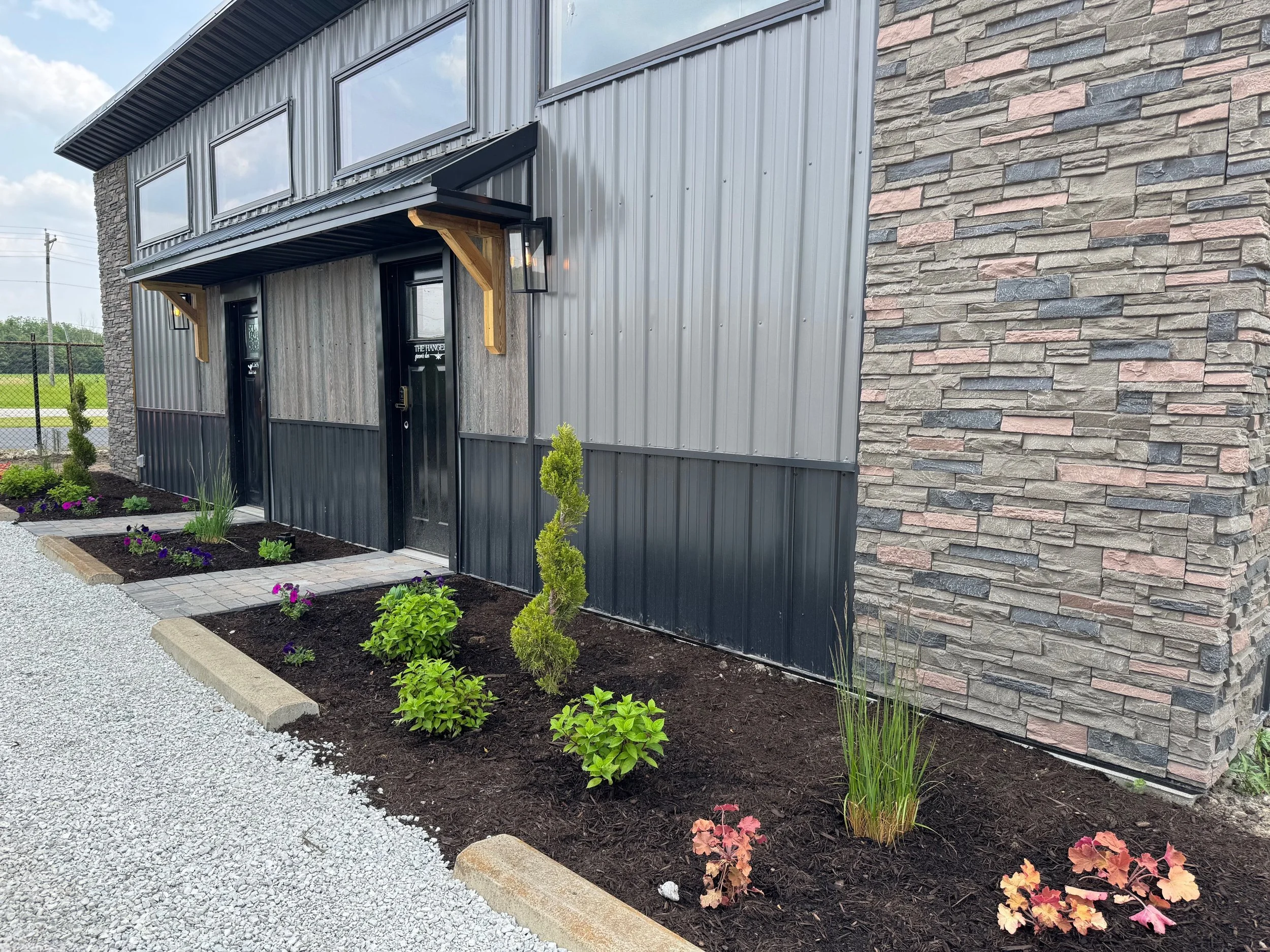 A modern building with a combination of stone, corrugated metal, and large windows. The entrance has a black door, outdoor lighting, and a small landscaped garden with young shrubs and flowers, bordered by a gravel pathway.