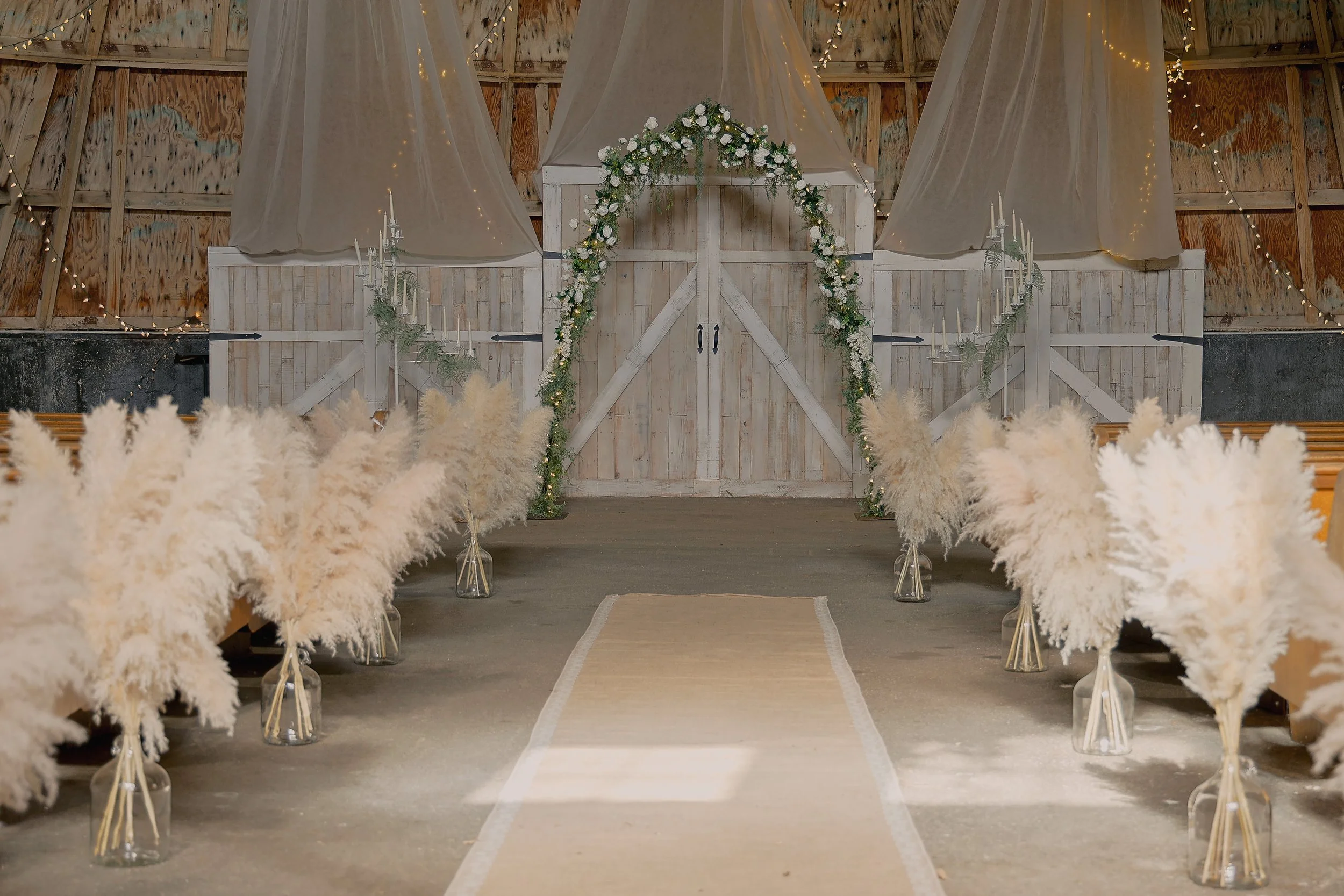 Wedding ceremony setup with an aisle, white pampas grass decorations in glass vases on either side, a floral arch with white flowers and green foliage at the altar, wooden barn walls, string lights, and cream curtains.