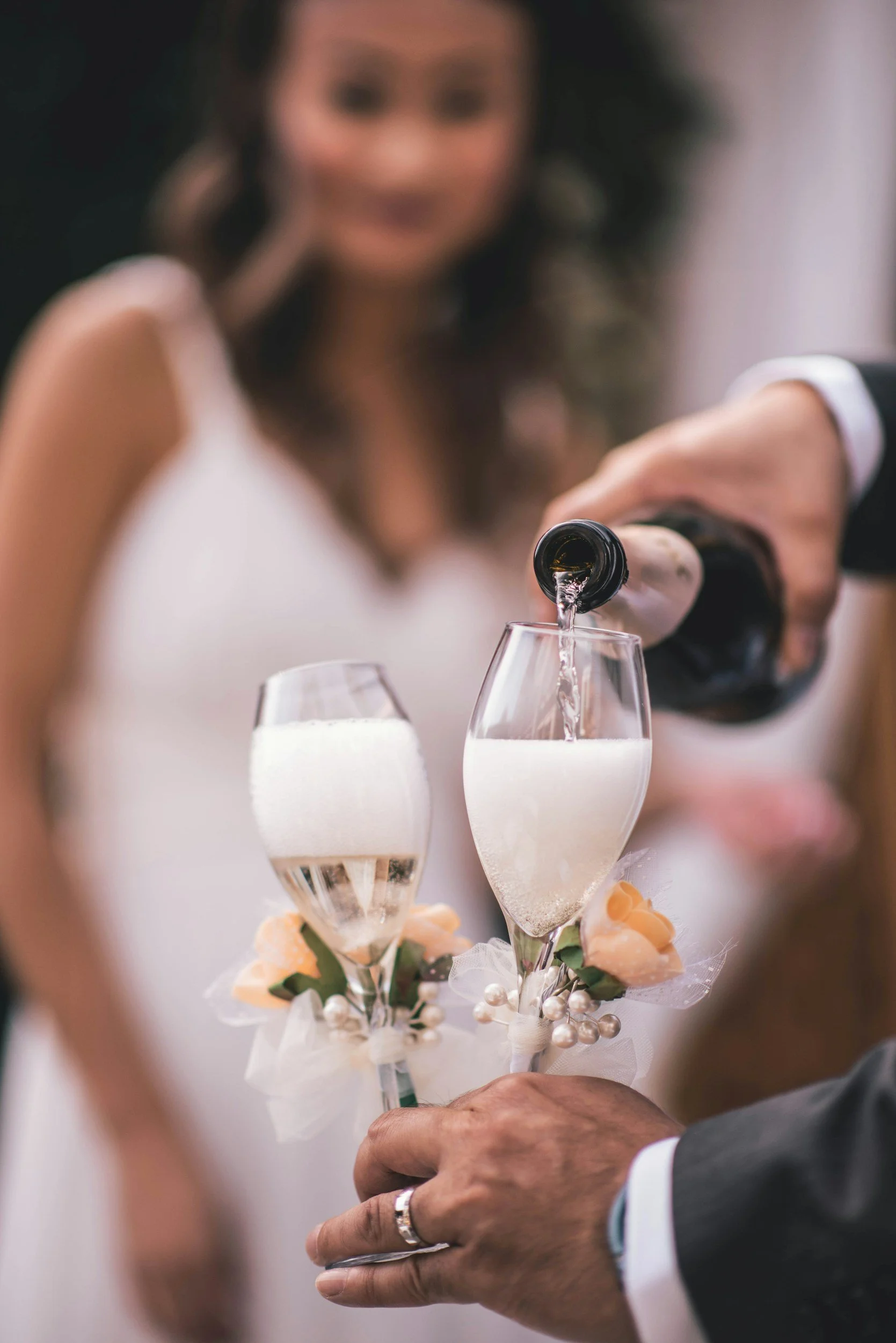 A couple is pouring champagne into glasses during their wedding ceremony, with the bride's sleeveless white dress and floral decorations visible.