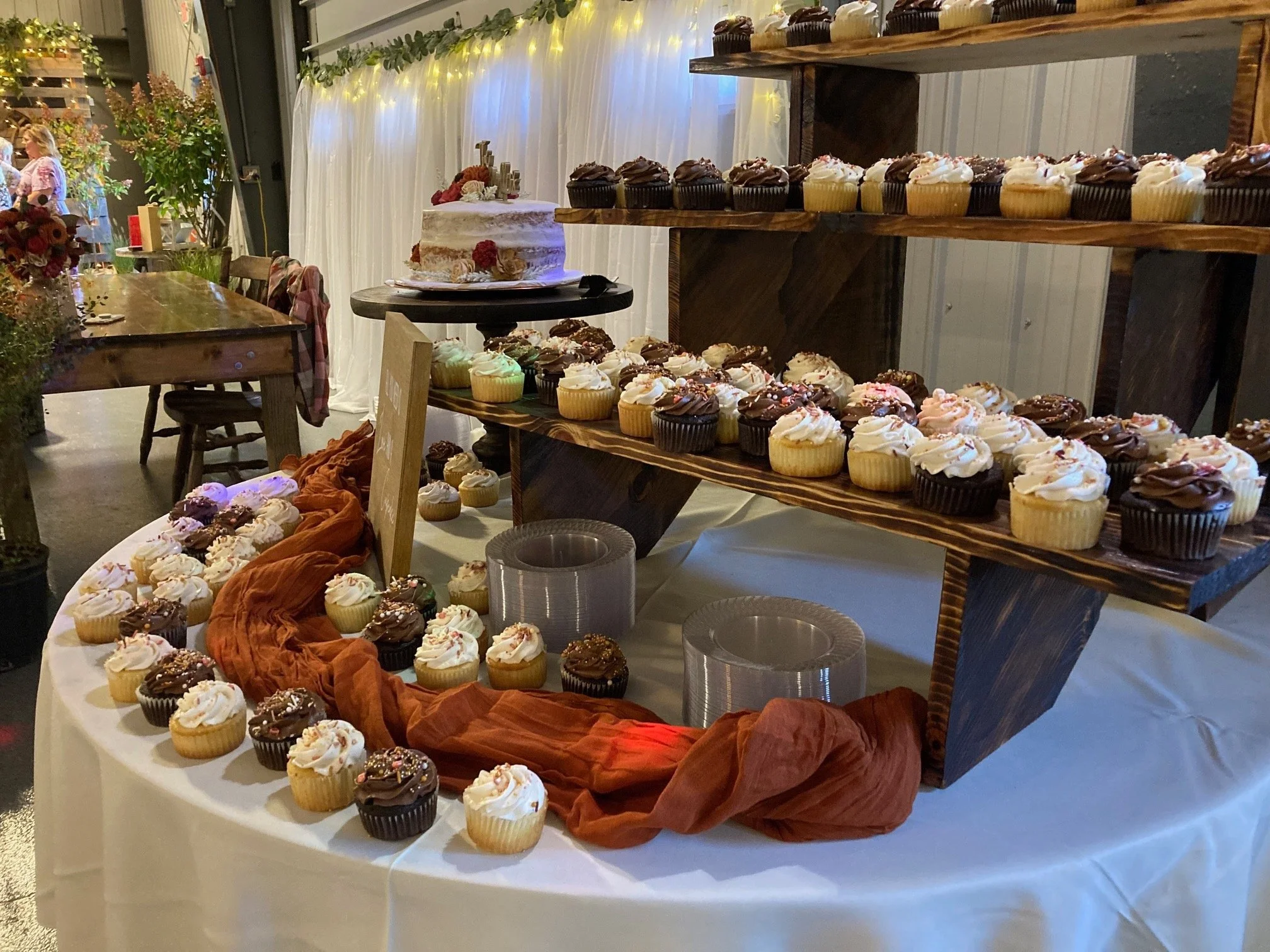 A dessert table with an assortment of cupcakes and a wedding cake at an indoor celebration with fairy lights and greenery in the background.