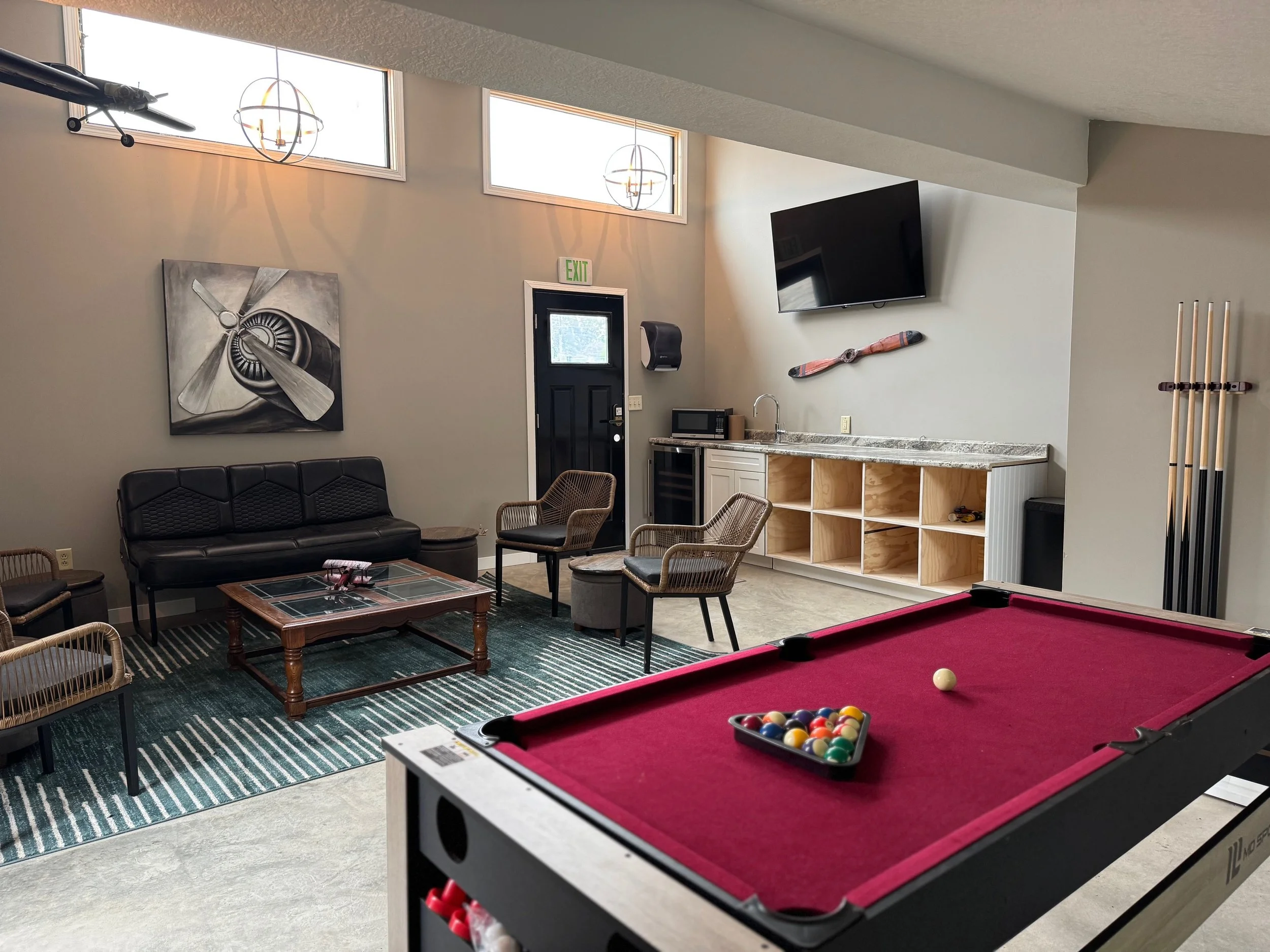 An indoor recreational room featuring a pool table with a red felt surface, billiard balls arranged in a triangle, and a cue stick holder on the wall. Other furniture includes a black leather sofa, wicker chairs, a coffee table, and a kitchenette with a microwave and sink. The room has high ceilings with two rectangular windows and modern light fixtures.