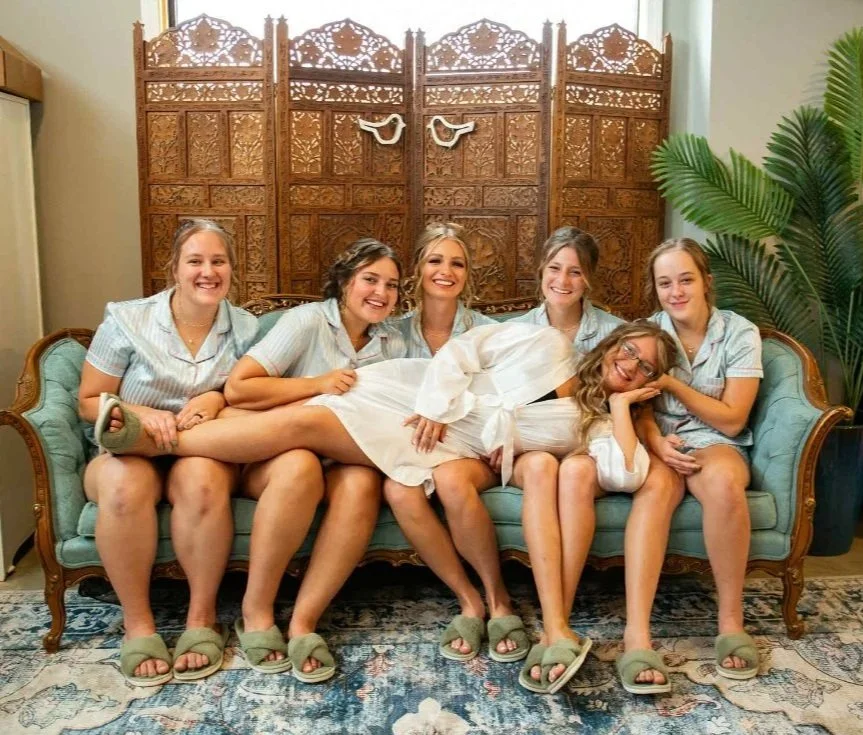 Six women wearing matching light blue pajamas and slippers sitting on a vintage teal sofa in front of a decorative wooden room divider, with a large green plant on the right side.