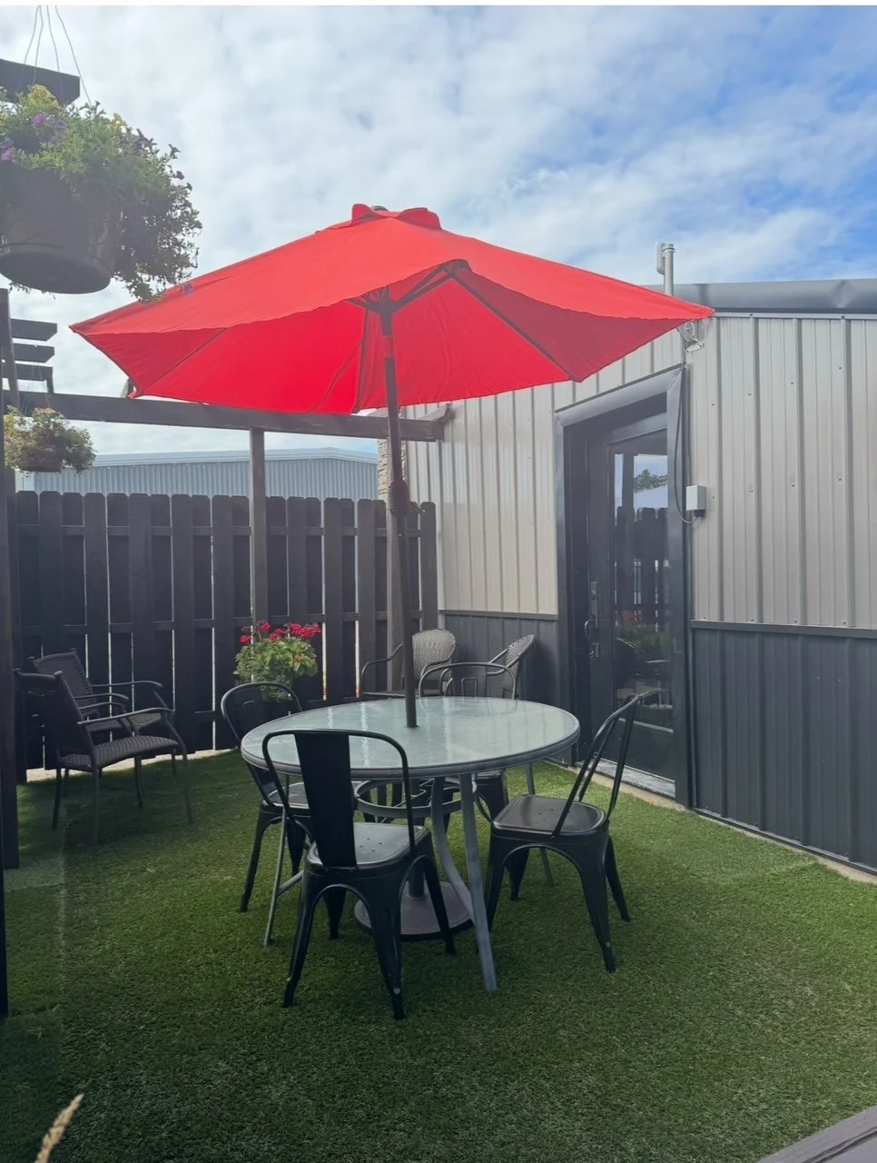 Outdoor patio with a round table, six black chairs, a red umbrella, a fence, potted flowers, and a building with a sliding door.