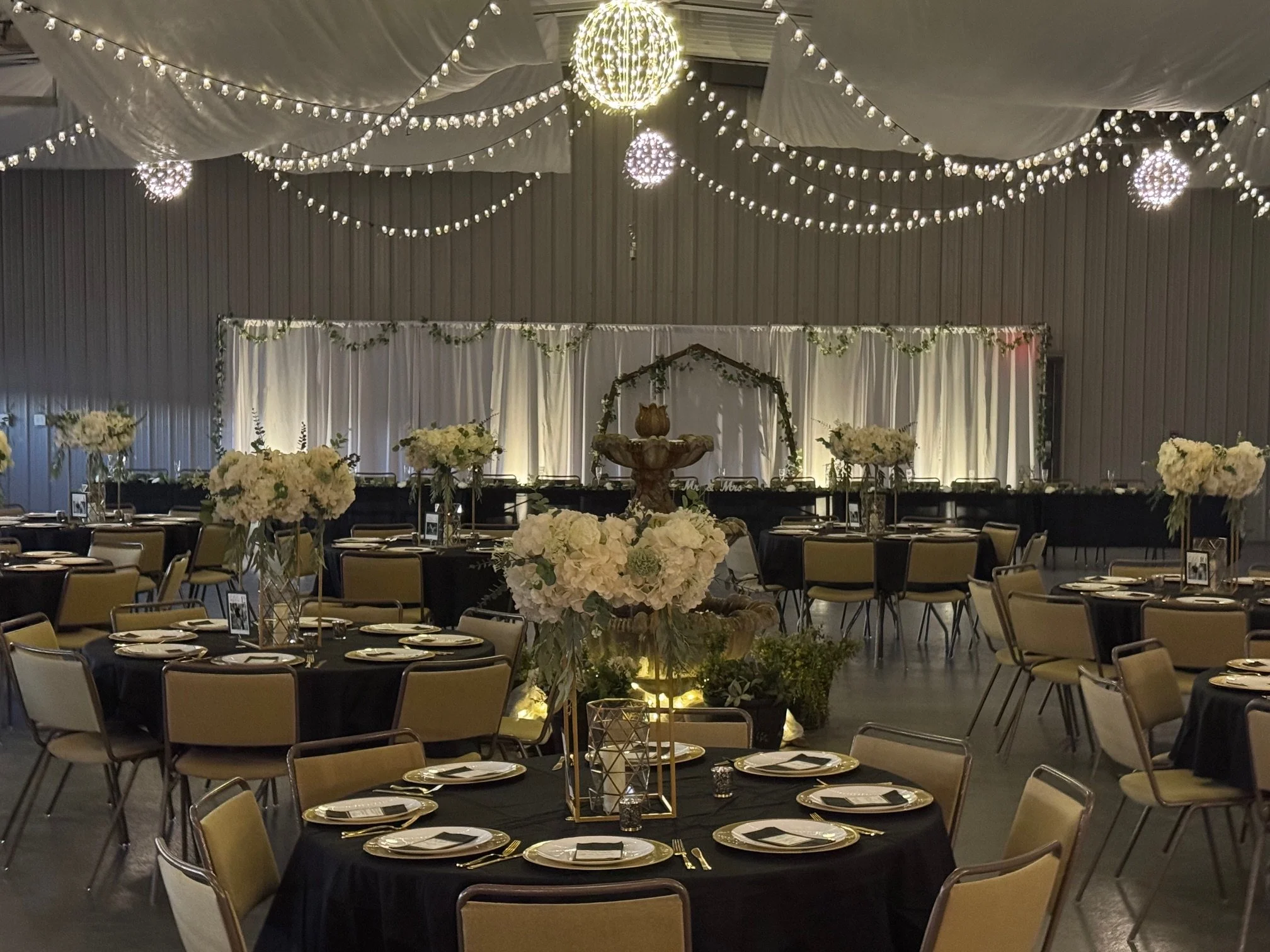 Elegant banquet hall decorated with string lights, draped fabric ceiling, and floral centerpieces on round tables with black tablecloths, set for a formal event.