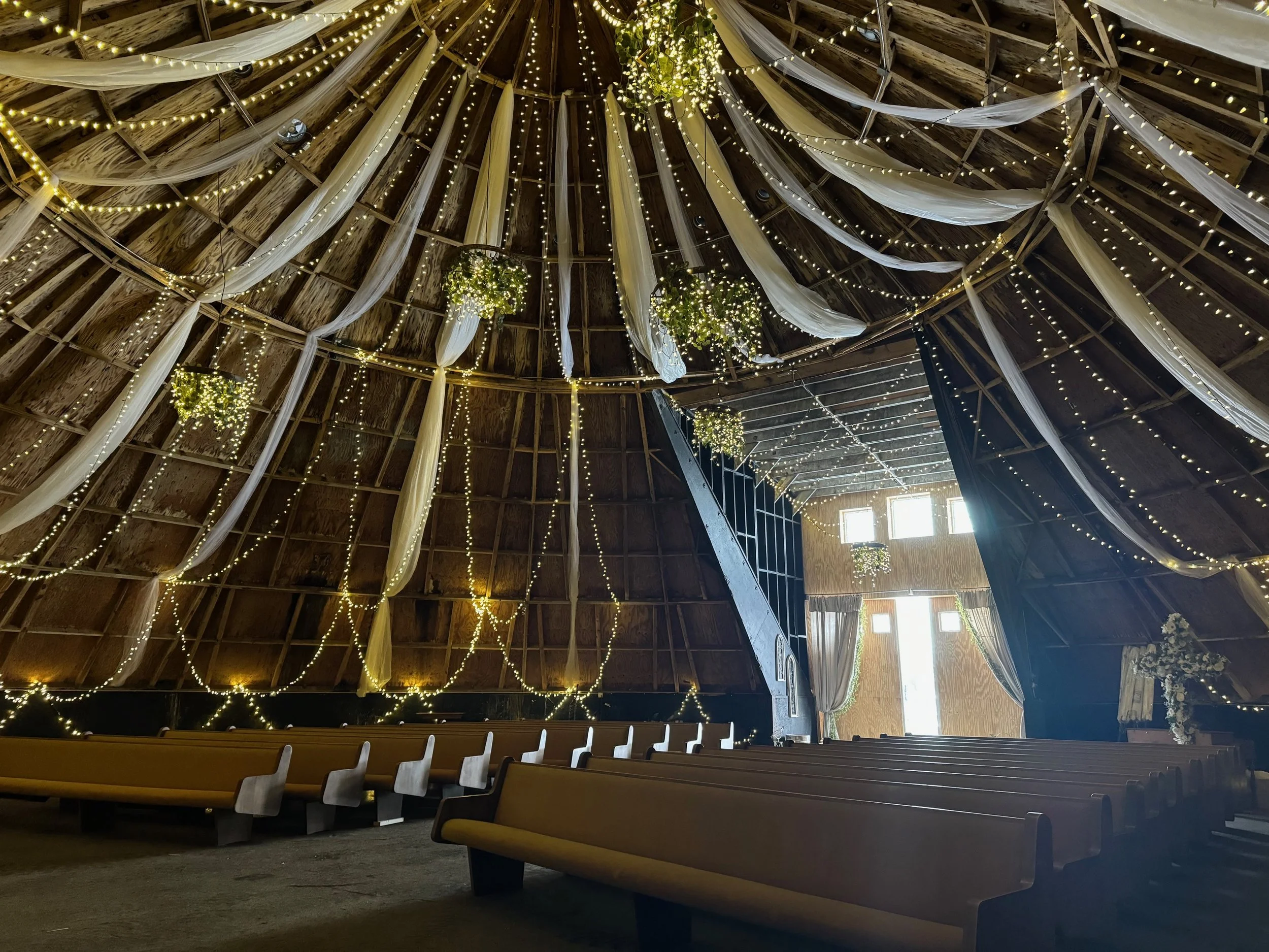 Interior of a rustic barn decorated for a wedding or special event, with string lights, hanging greenery, and white drapery.