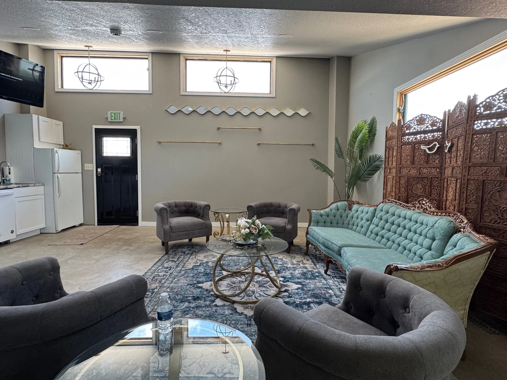 Living room with vintage teal sofa, gray armchairs, small round glass table with floral centerpiece, patterned rug, decorative wooden screen, large window, and plants.